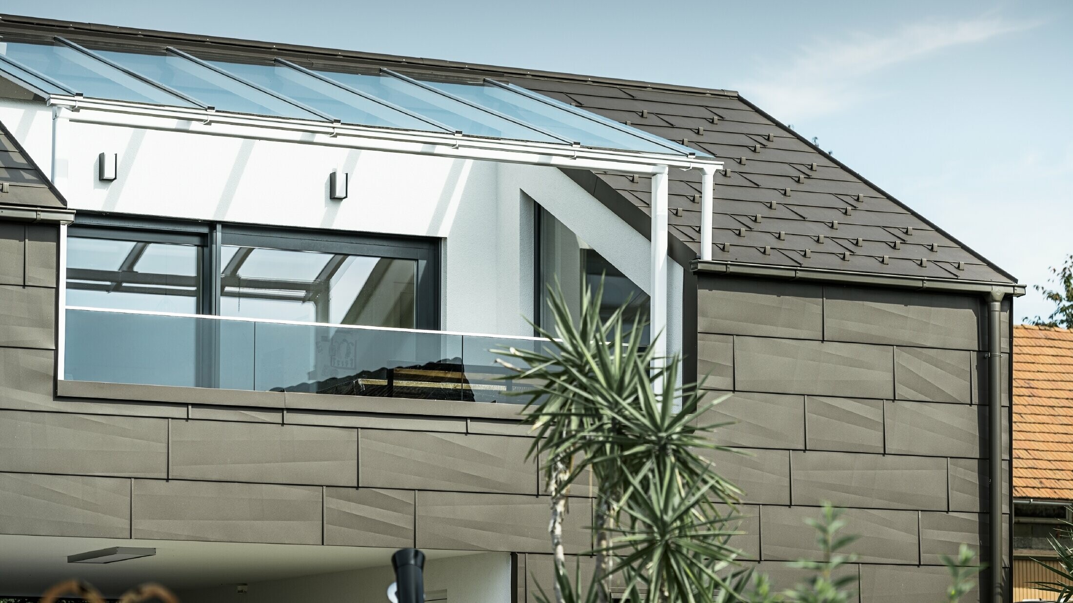 Extension avec terrasse de toit revêtue du système complet PREFA ; pour le toit et la façade, le choix s'est porté sur le panneau de toiture et de façade FX.12 PREFA. Pour l'écoulement des eaux de pluie, le choix s'est également porté sur la gouttière carrée PREFA avec tuyau de descente PREFA et accessoires complets en brun.
