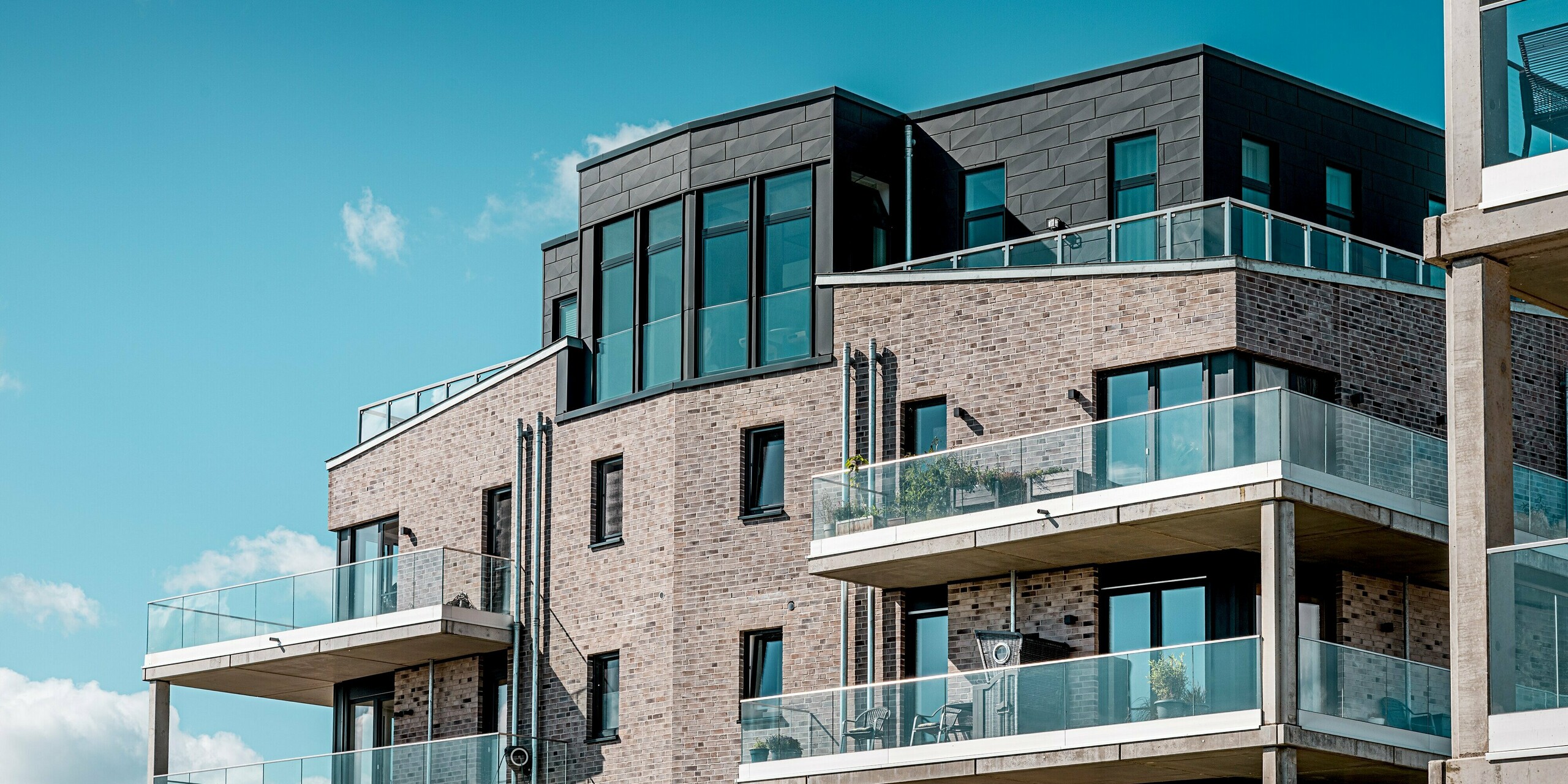 Die moderne Wohnanlage „Hafen zur Freiheit“ zeigt im oberen Bildausschnitt eindrucksvoll die markante Fassadengestaltung mit der Kombination aus hellem Klinker und elegantem PREFA Siding.X in P.10 Anthrazit. Besonders auffällig sind die kraftvoll strukturierten Aluminiumflächen rund um das Staffelgeschoss, die dem Gebäude eine dynamische, fast skulpturale Wirkung verleihen. Die großzügigen Glasbalkone reflektieren das Licht und betonen die hochwertige Architektur. Die langlebigen, wetterfesten PREFA Aluminiumpaneele sorgen zudem für optimalen Schutz in der exponierten Küstenlage und steigern die Wertbeständigkeit.