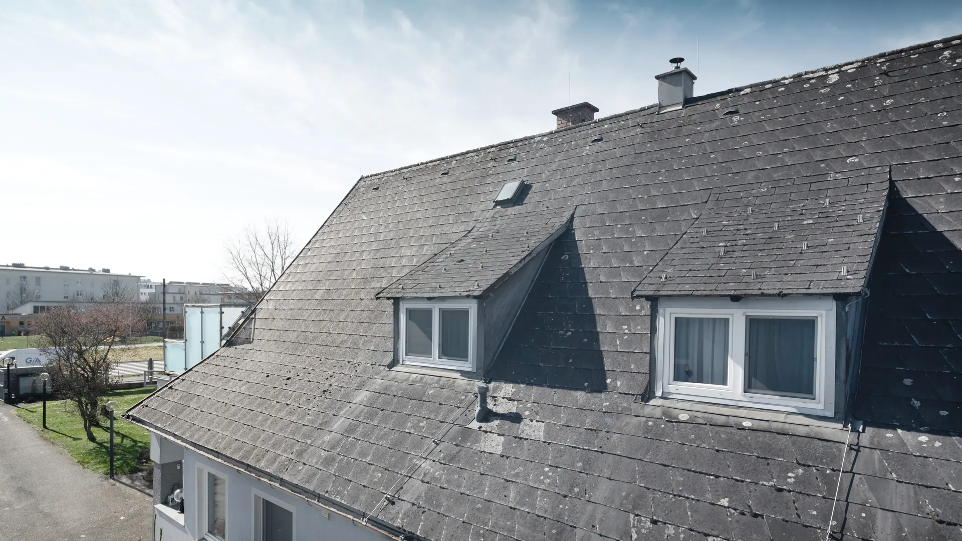 Detailaufnahme eines alten Eternit-Dachs mit sichtbaren Abnutzungsspuren und Flechtenbewuchs. Das Dach hat zwei Gauben, die sichtbar sind. Aufnahme vor der Dachsanierung.