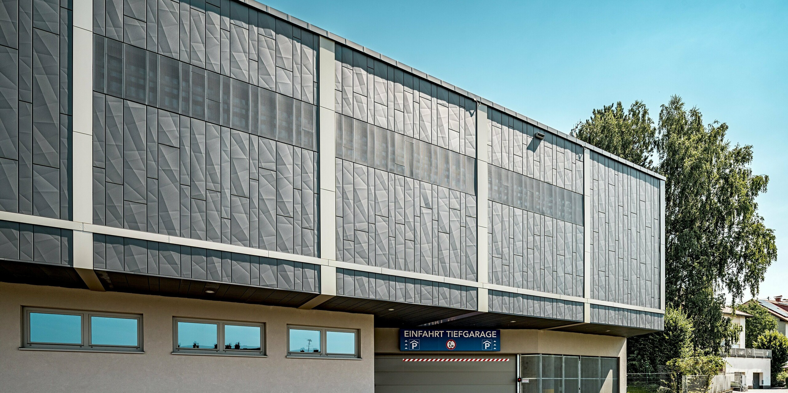 Das Bild zeigt die Zufahrt zur Tiefgarage des Einkaufszentrums in Rohrbach. Das Gebäude wurde mit PREFA Siding X-Paneelen in Anthrazit verkleidet. Besonders auffällig sind die geometrisch strukturierten Fassadenelemente, die dem Gebäude eine moderne Ausstrahlung verleihen und den Bereich der Tiefgarageneinfahrt perfekt abrunden. Die klare Architektur wird durch die präzise Platzierung der Fenster und perforierten Sidings zusätzlich betont.