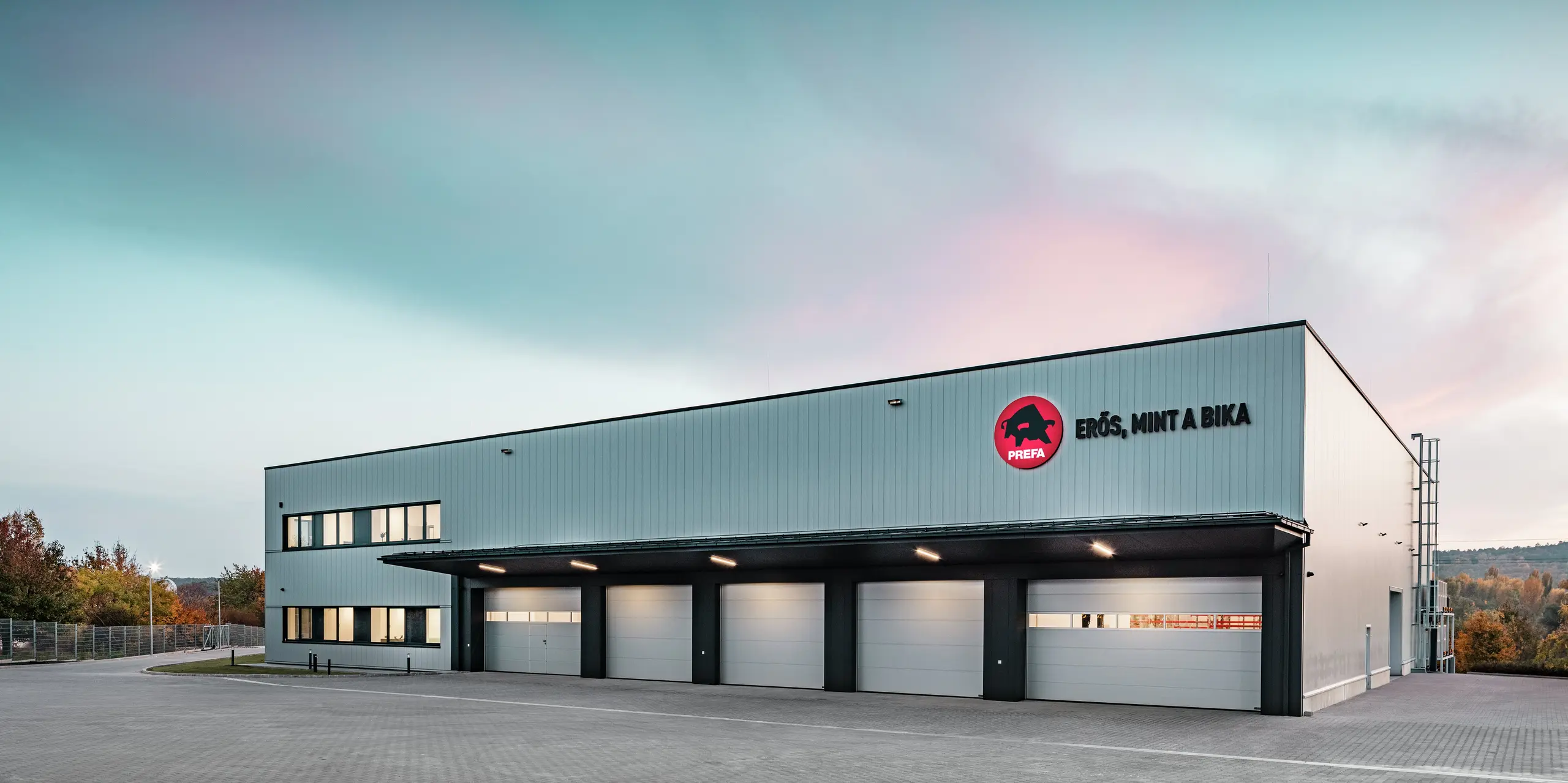 La vaste zone de stockage et de logistique du site PREFA à Törökbálint (Hongrie) impressionne par son architecture moderne et fonctionnelle. La façade en Sidings PREFA en argent métallisé confère au bâtiment une surface élégante, durable et résistante aux intempéries. Le logo PREFA frappant avec le slogan « Erős, mint a bika » (« Robuste comme un taureau ») souligne la qualité et la robustesse des produits en aluminium.