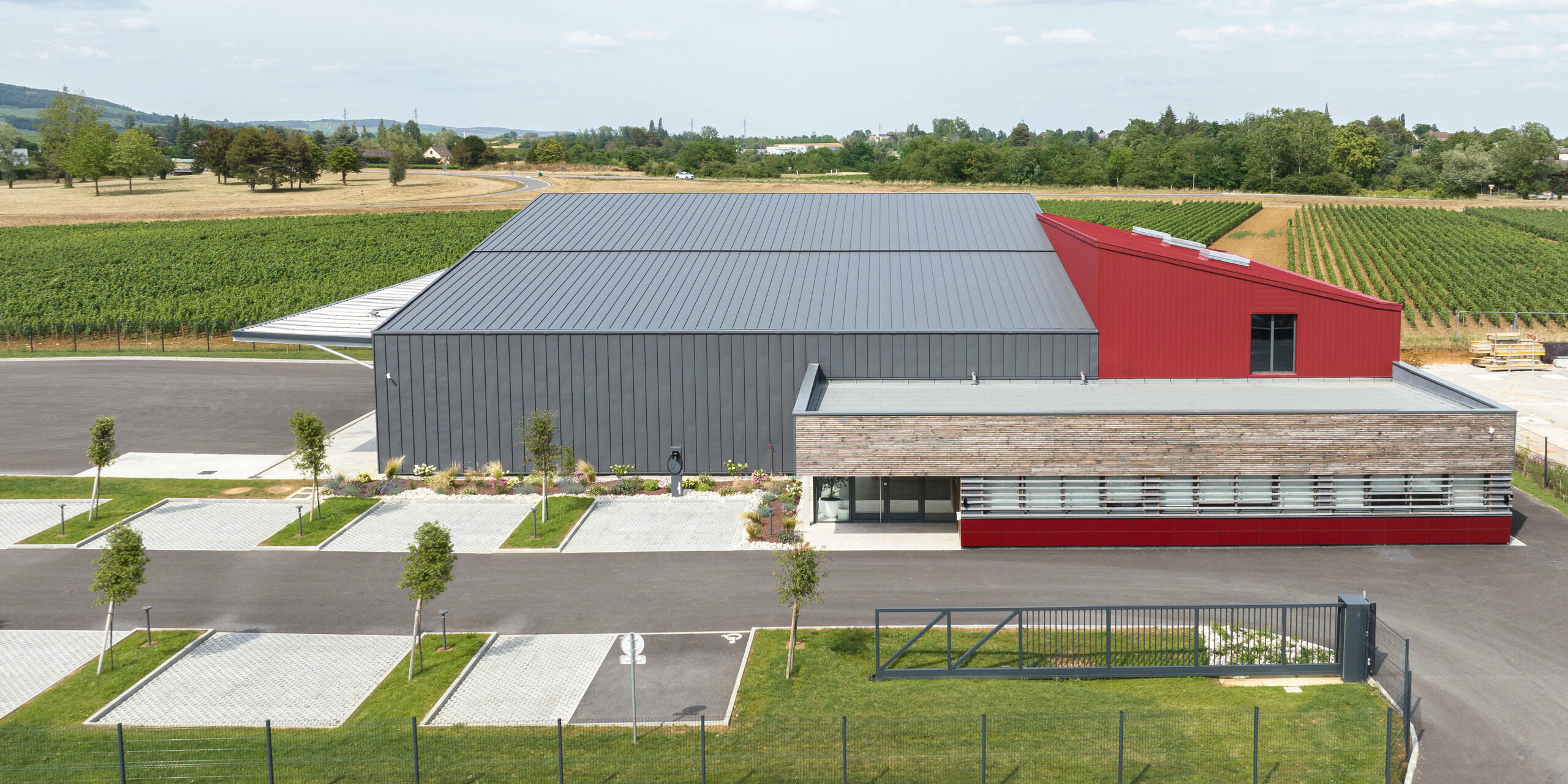 Ein großes Industriegebäude mit PREFALZ in Dunkelgrau am Dach und der Fassade. Als farblicher Akzent wurden Teile des Gebäudes mit PREFALZ bzw. PREFA Sidings in P.10 Oxydrot verkleidet. Das Gebäude steht vor einer Landschaft mit Feldern im Hintergrund. Im vorderen Bildrand sind Parkplätze mit jungen Bäumen und einer ordentlichen Bepflanzung zu sehen.