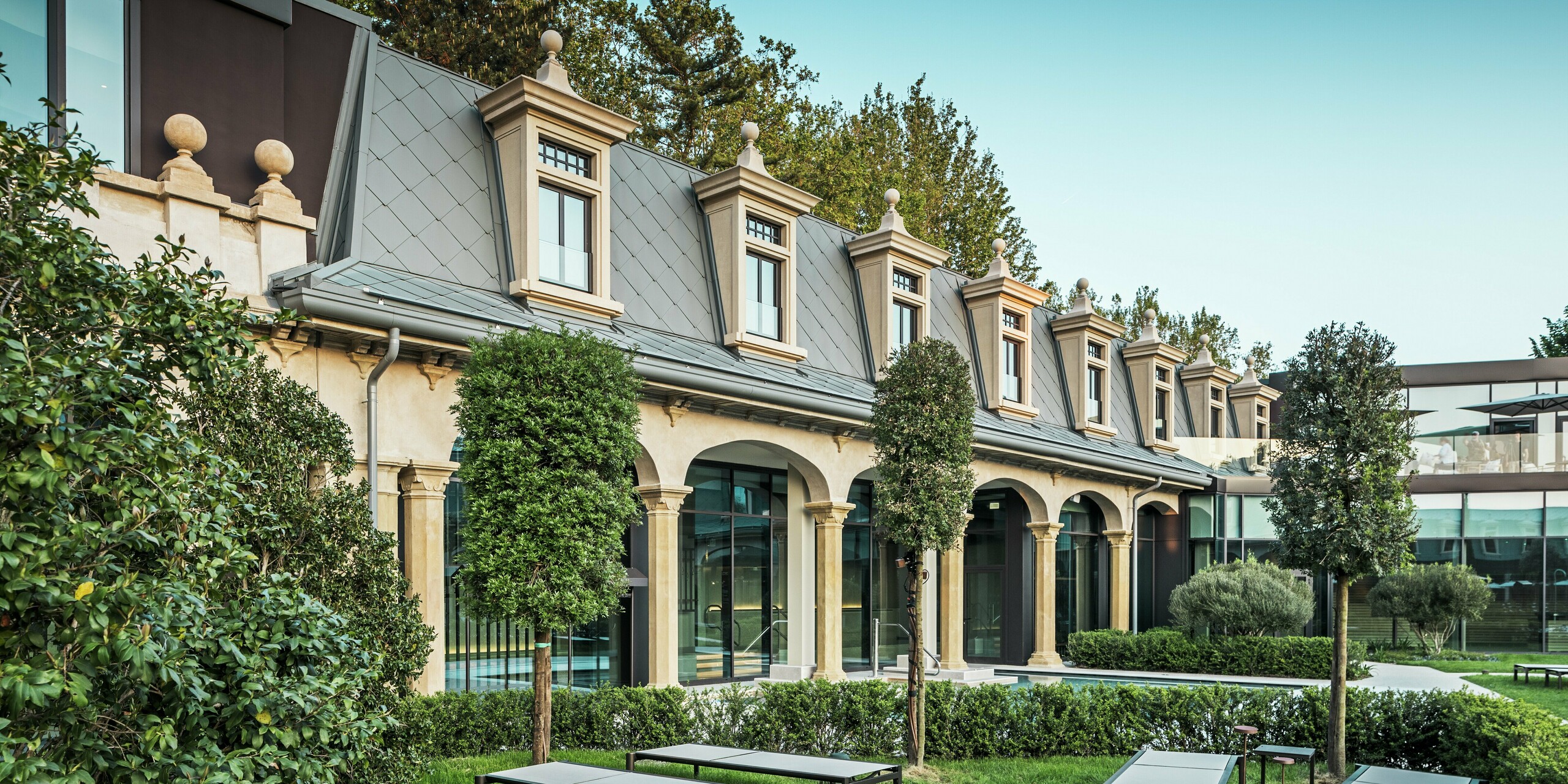 Vue détaillée de la toiture rénovée avec style des thermes « De Montel – Terme Milano », avec ses losanges PREFA 44 × 44 en P.10 gris souris. Les lucarnes, les finitions des fenêtres et les corniches, réalisées avec précision, soulignent l'élégance historique de cet ensemble Art nouveau. Au premier plan, un jardin paisible avec des chaises longues modernes, des haies taillées géométriquement et de beaux arbres – un lieu de détente à la profondeur architecturale remarquable.