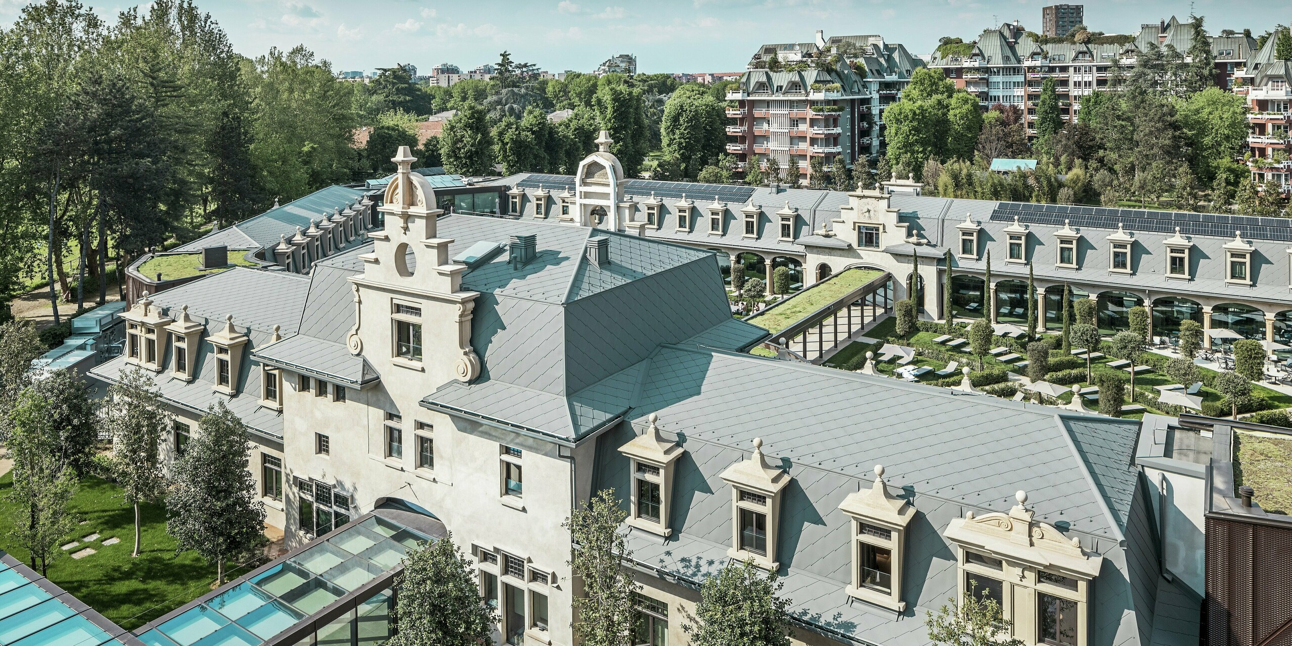 Vue d'ensemble de l'impressionnant complexe « De Montel – Terme Milano », alliant architecture Art nouveau classée et ambiance bien-être moderne. La toiture généreuse en losanges PREFA 44 × 44 P.10 gris souris confère au complexe une élégante uniformité. Les modules photovoltaïques intégrés, les toitures végétalisées et les liaisons vitrées entre les bâtiments témoignent de l'engagement en faveur du développement durable et du design contemporain en contexte urbain.