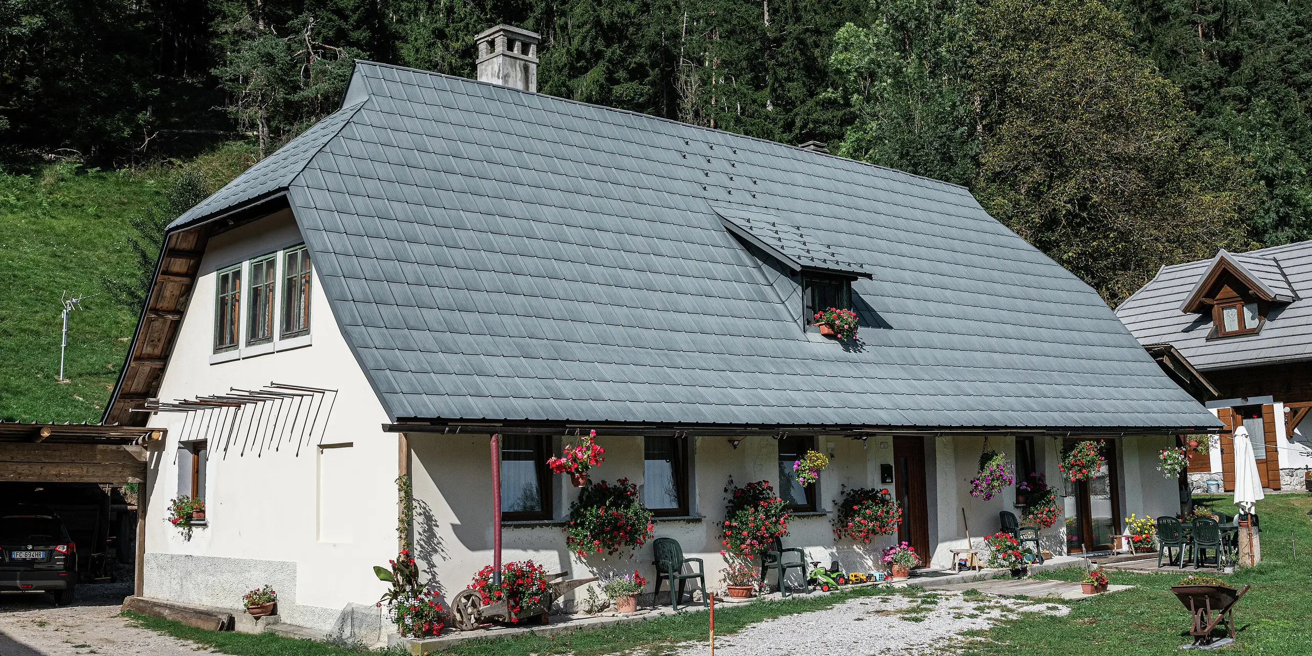 Une maison familiale idyllique à Tarvisio, en Italie, est recouverte de tuiles PREFA durables depuis 1972. Le grand toit en croupe en aluminium résistant aux intempéries protège la maison des conditions météorologiques alpines depuis des décennies. La façade est décorée de nombreuses fleurs, créant une atmosphère accueillante. Une petite terrasse avec des sièges vous invite à vous attarder. À l'arrière-plan s'étend une forêt dense, qui intègre harmonieusement la maison dans la nature. Sur la gauche, vous pouvez voir une allée couverte avec une voiture garée, tandis qu'à droite, une autre charmante maison en bois complète le tableau.