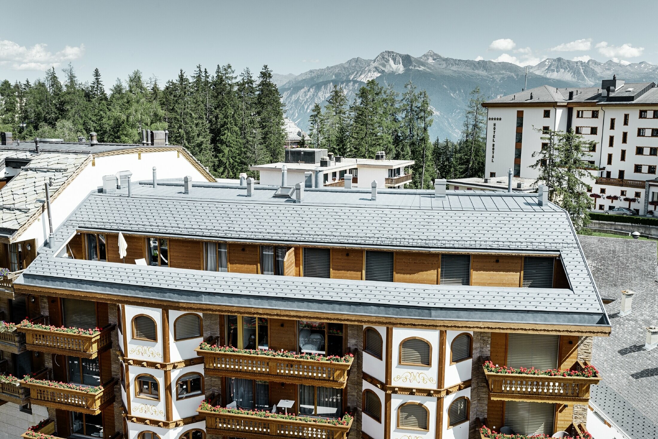 Immeuble d’habitation de Crans-Montana (Suisse) avec les Alpes en arrière-plan — Façade intégrant de nombreux éléments en bois et dont la toiture a été réalisée avec des bardeaux en aluminium PREFA de couleur gris pierre