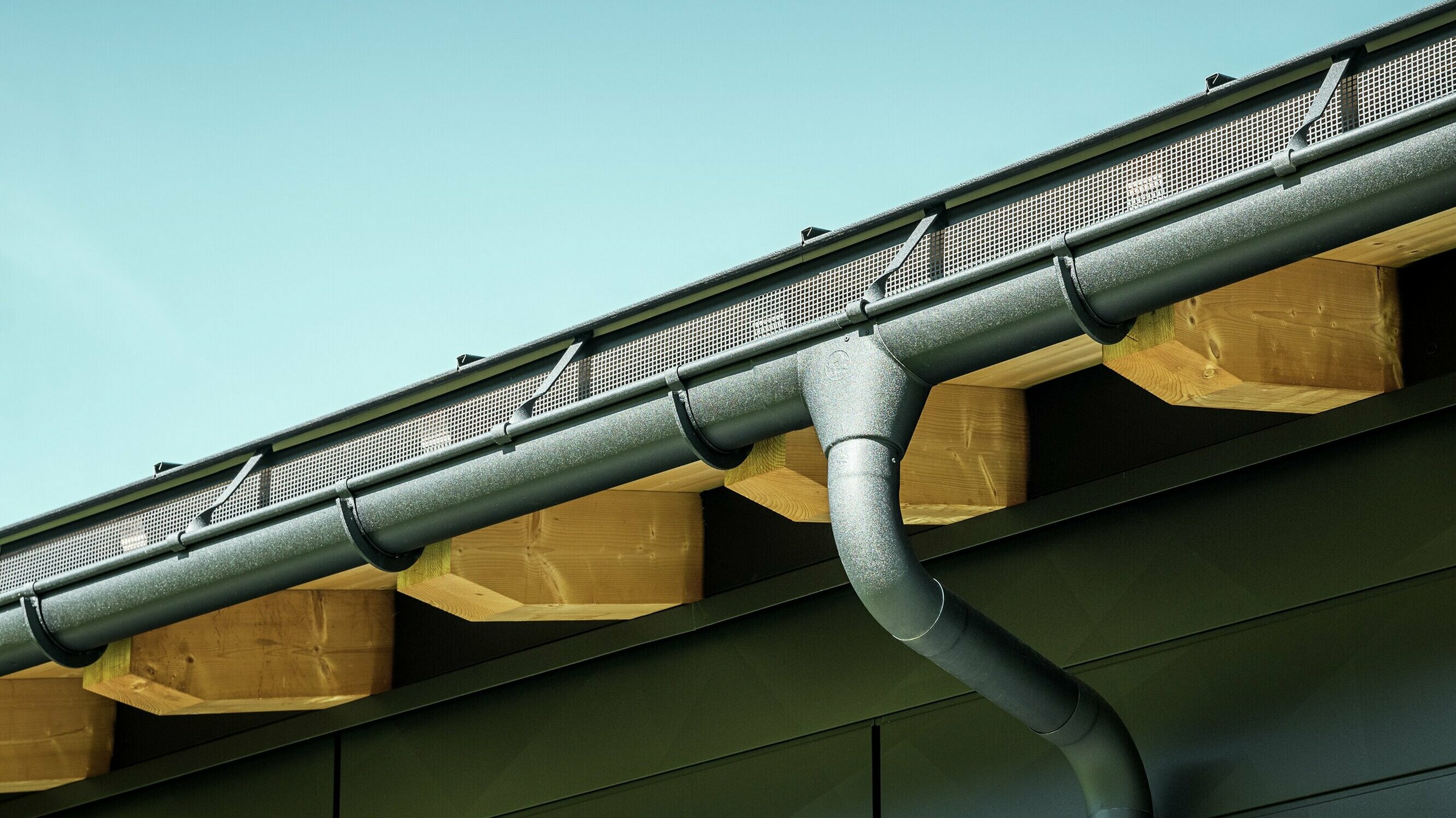 Close-up van de gevel van een moderne eengezinswoning in Ascharina, Zwitserland. De zijkanten van het gebouw zijn bekleed met de opvallende PREFA Siding.X gevelpanelen in P.10 zwart. De PREFA dakafvoersystemen met mastgoot en afvoerbuis zijn qua kleur op elkaar afgestemd.