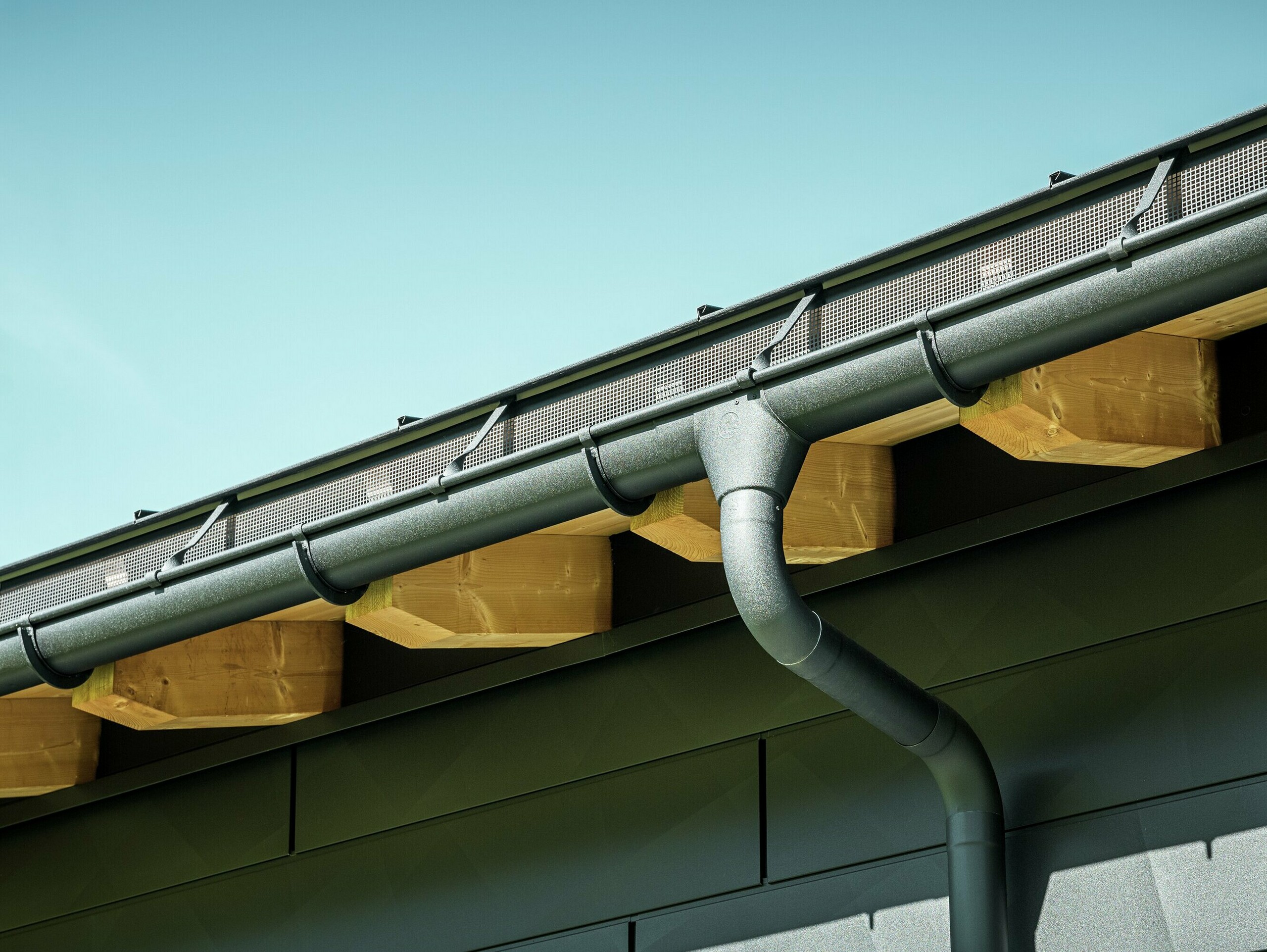 PREFA mastgoot met trechteruitloop en oversteek in P.10 zwart Close-up van de gevel van een moderne eengezinswoning in Ascharina, Zwitserland. De zijkanten van het gebouw zijn bekleed met de opvallende PREFA Siding.X gevelpanelen in P.10 zwart. De PREFA dakafvoersystemen met mastgoot en afvoerbuis zijn qua kleur op elkaar afgestemd.