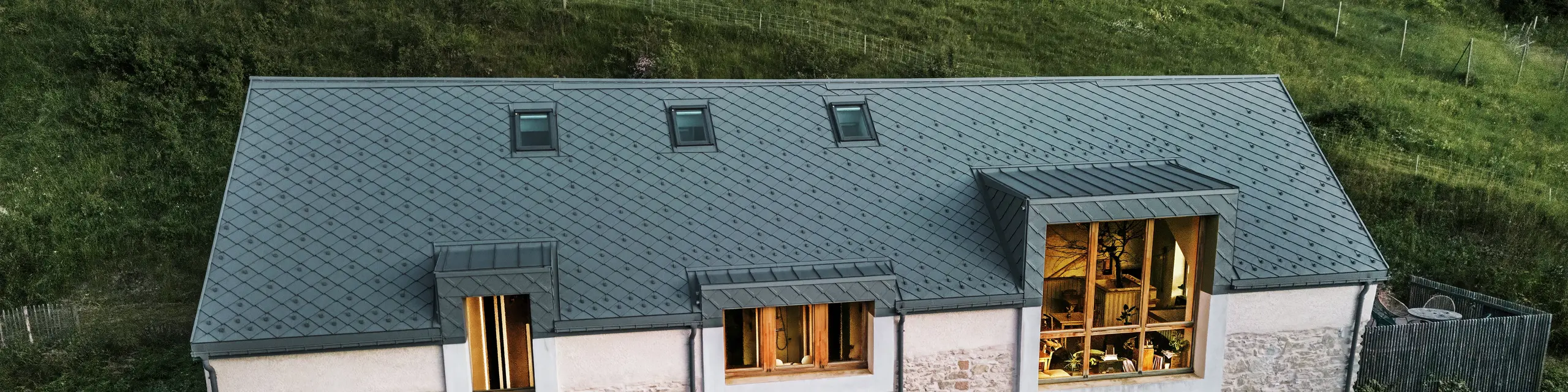Abendansicht der renovierten Scheune „La Grange à Gaby“ in Engins, Frankreich. Das Gebäude vereint traditionelles Steinmauerwerk mit einem modernen PREFA-Aluminiumdach mit 29 × 29 cm großen Rauten. Große Fenster und helle Innenräume unterstreichen die behagliche Atmosphäre des neuen Kulturzentrums.