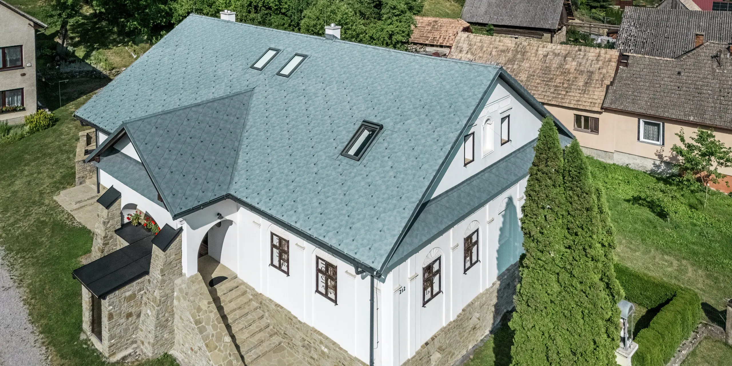 Das Bild zeigt das sanierte Handwerkerhaus in Klin, Slowakei, von der Vogelperspektive. Das schlichte Satteldach mit einer Gaube über dem Eingang wurde mit PREFA Dachrauten 44 × 44 in P.10 Steingrau eingedeckt. Die Dachfläche wird von symmetrischen Rauten geprägt, die dem Gebäude eine moderne, zugleich elegante Ausstrahlung verleihen. Die integrierten Dachfenster sorgen für zusätzliches Tageslicht und betonen die Funktionalität des Designs. Das Aluminiumdach bietet zahlreiche Vorteile: Es ist leicht, langlebig, korrosionsbeständig und ideal für die Sanierung von historischen Gebäuden, da es die Dachlast minimiert und gleichzeitig eine hohe Widerstandsfähigkeit gegenüber Witterungseinflüssen bietet.