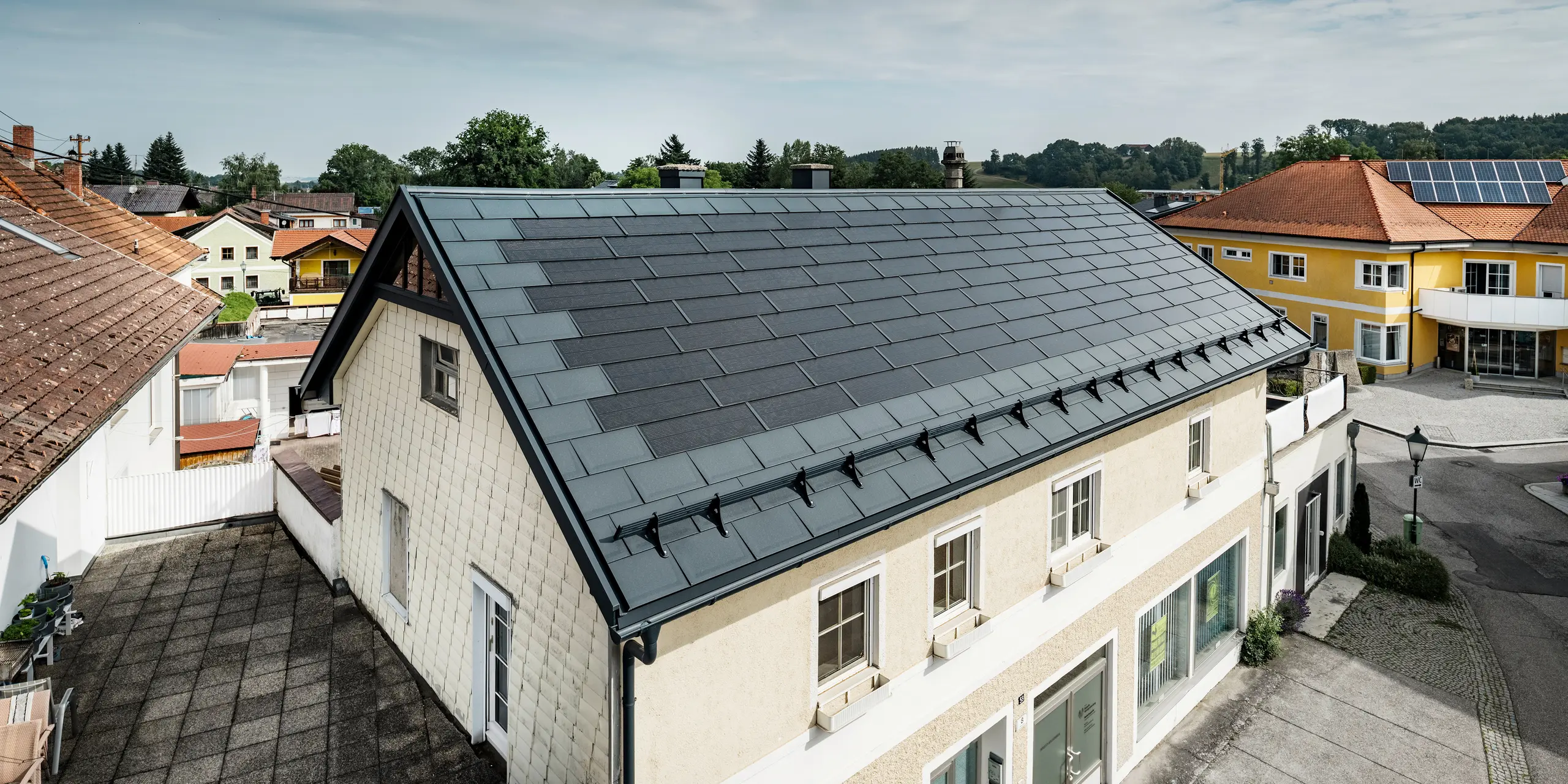 Vue du dessus d’une maison familiale à Mettmach, en Autriche. Le bâtiment le plus ancien a été rénové avec des tuiles solaires PREFA de couleur anthracite. Les systèmes de protection contre la neige et les lignes claires des panneaux solaires soulignent la finition soignée des produits en aluminium PREFA. Le toit de tuiles orange d’un immeuble voisin contraste en arrière-plan. Les modules photovoltaïques sont installés sur le toit de la maison individuelle voisine dans une solution de toit classique, qui montre la diversité des conceptions de toit économes en énergie.