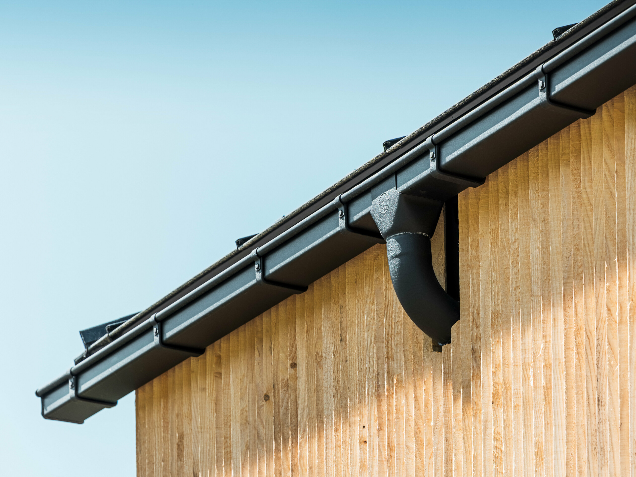 Hauseck mit der PREFA Kastenrinne in Schwarz, mit Rinnenkasten, befestigt mit den PREFA Rinnenhaken, die Fassade ist mit Holz verkleidet..