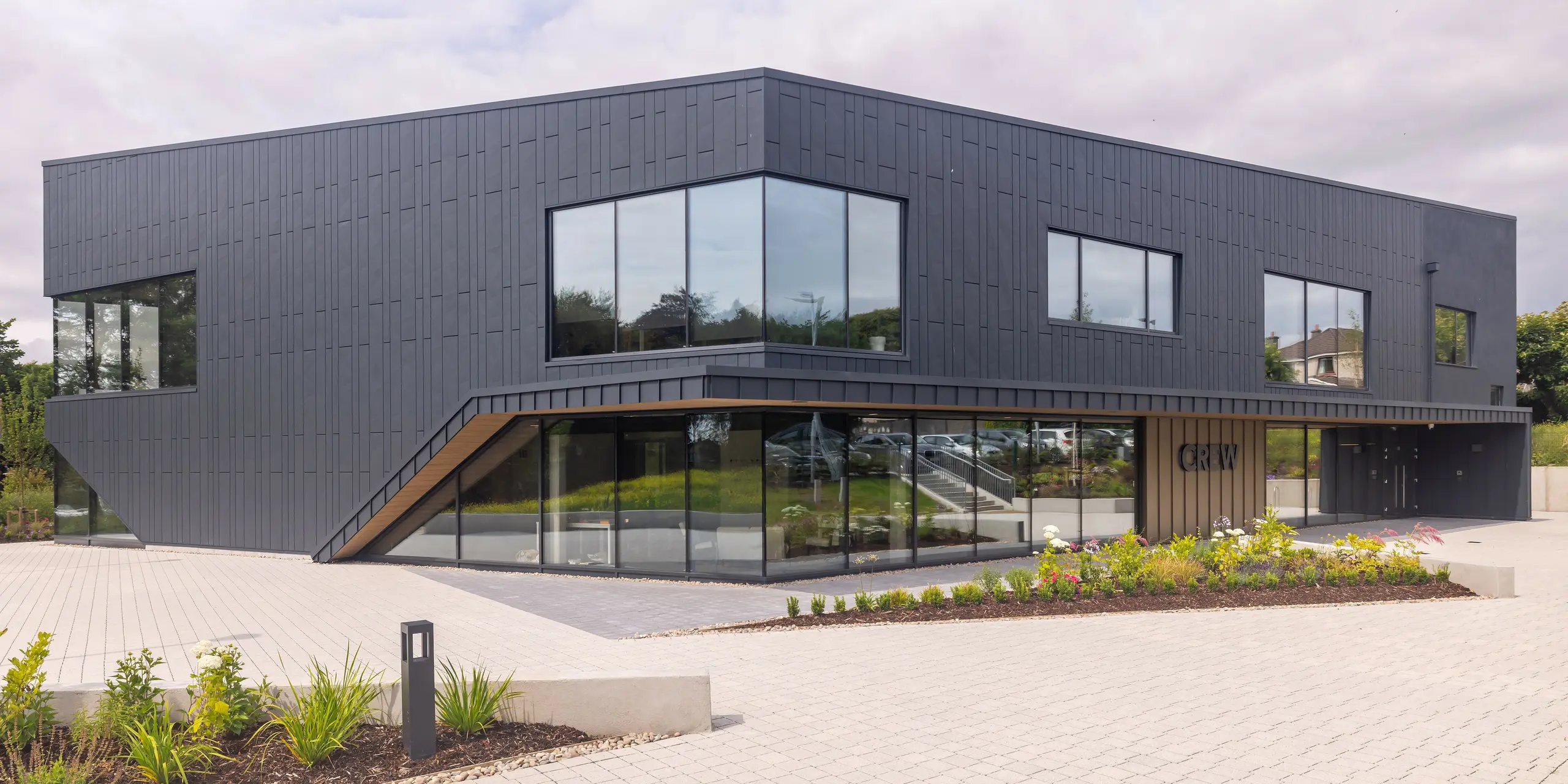 Frontalansicht des CREW-Gebäudes in Galway, Irland, mit moderner PREFA Aluminiumfassade aus Siding.X und PREFALZ in P.10 Anthrazit. Die schrägen Linien und großen Fensterflächen prägen das architektonisch anspruchsvolle Design des Gebäudes. Die anthrazitfarbene Aluminiumverkleidung in Kombination verleiht dem Gebäude eine elegante und zeitgemäße Ästhetik, die durch die gepflegte Landschaftsgestaltung ergänzt wird.