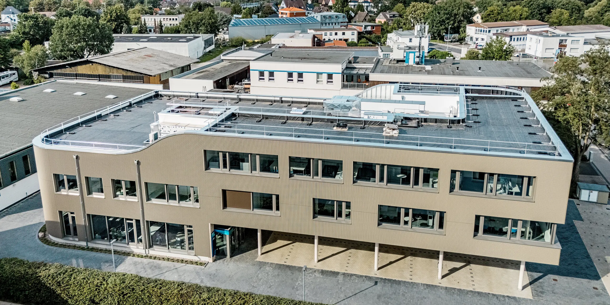 L'image présente le bâtiment Festo à Langenhagen, vu du ciel. Sa façade remarquable, composée de profils extrudés triangle PREFA à la finition bronze, lui confère une allure moderne et dynamique. La courbe de la partie supérieure de la façade est particulièrement visible et lui donne une forme élégante et contemporaine. Les larges fenêtres et la structure apparente du mur extérieur offrent une harmonieuse combinaison de fonctionnalité et d'esthétique. L'utilisation des produits PREFA garantit une longue durée de vie et une excellente résistance aux intempéries. La précision de l'exécution et la qualité des matériaux font de ce bâtiment un exemple exceptionnel d'architecture moderne.