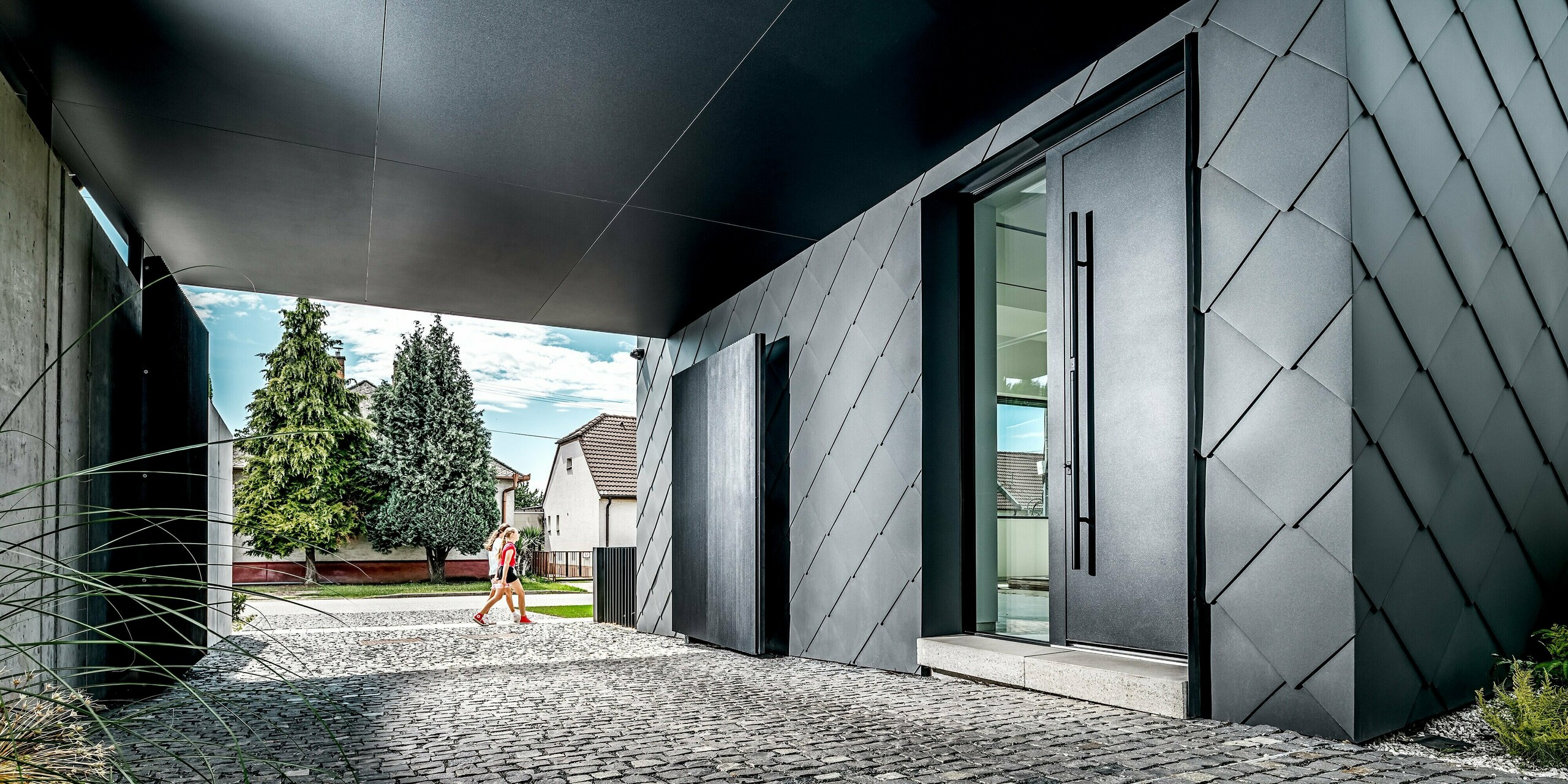 Dieses Bild zeigt den Eingangs- und Durchfahrtsbereich eines modernen Einfamilienhauses in Trnava-Modranka, dessen markante Fassade vollständig mit PREFA Wandrauten 44 × 44 in P.10 Dunkelgrau verkleidet ist. Die präzise Rautenstruktur verleiht dem Gebäude eine elegante, futuristische Wirkung und betont die klare Linienführung der Architektur. Die widerstandsfähigen Aluminiumprodukte sorgen für dauerhaften Schutz vor Witterung und eine hochwertige, langlebige Optik. Im Hintergrund setzt das begrünte Wohnumfeld einen natürlichen Kontrast zur dunklen Gebäudehülle.