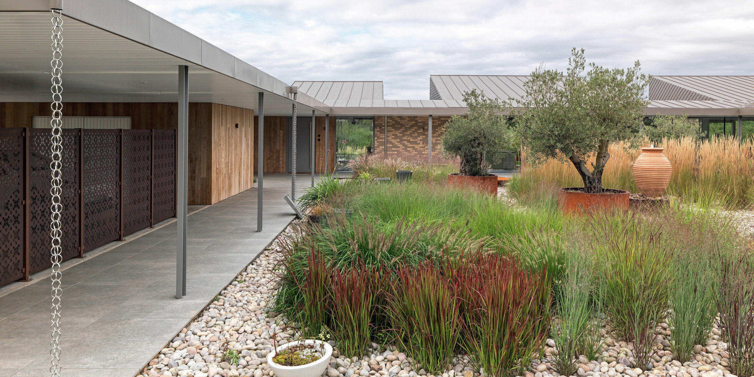 Vue détaillée d'une maison moderne du West Sussex avec toit plat en aluminium PREFALZ couleur  P.10bronze. On y découvre une terrasse couverte et clairement structurée, avec des colonnes fines, des chaînes d'évacuation des eaux pluviales intégrées avec style et un jardin varié composé de graminées ornementales, d'oliviers et de pierres naturelles. Les toitures durables en aluminium PREFA soulignent les lignes modernes et s'intègrent harmonieusement au concept architectural durable.