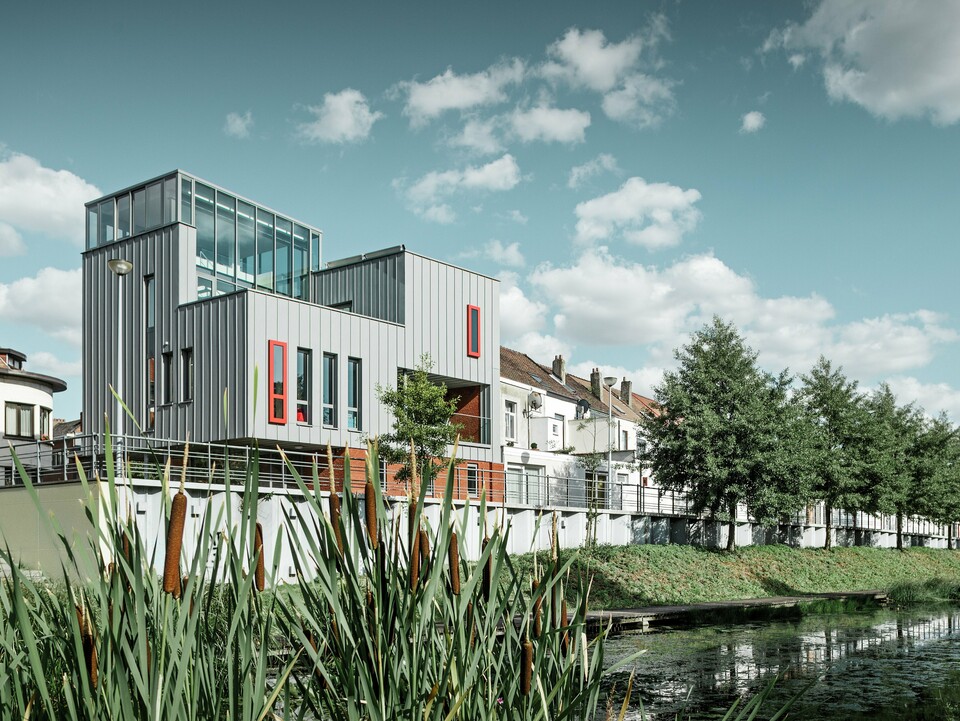 Architektonisches Highlight am Wasser: Das moderne Wohn- und Bürogebäude besticht durch eine PREFALZ Aluminiumfassade in Patinagrau mit vertikaler Stehfalzstruktur. Farblich abgesetzte Fenster in Rot setzen gezielte Akzente. Die Kombination aus Glas, Aluminium und rotem Klinker schafft ein harmonisches Gesamtbild – witterungsbeständig, pflegeleicht und langlebig.