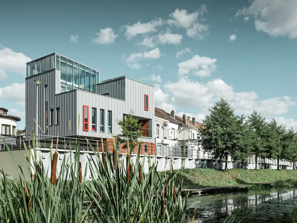 Architektonisches Highlight am Wasser: Das moderne Wohn- und Bürogebäude besticht durch eine PREFALZ Aluminiumfassade in Patinagrau mit vertikaler Stehfalzstruktur. Farblich abgesetzte Fenster in Rot setzen gezielte Akzente. Die Kombination aus Glas, Aluminium und rotem Klinker schafft ein harmonisches Gesamtbild – witterungsbeständig, pflegeleicht und langlebig.