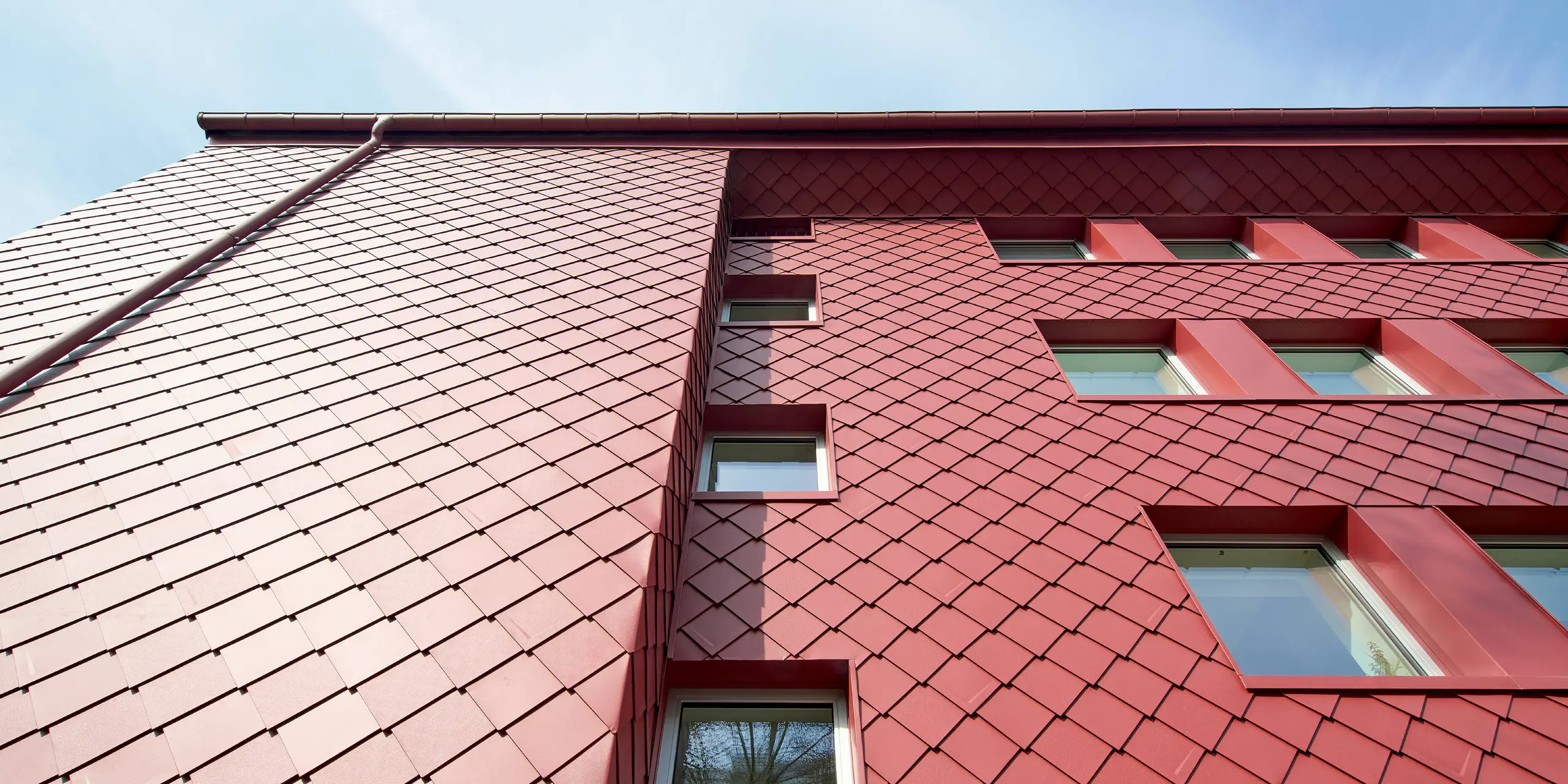 Spektakuläre Perspektive auf die PREFA Aluminiumfassade des Sanierungsprojekts „Energiesprong 2426“ in Köln-Zollstock. Die Wandrauten 29 × 29 in P.10 Oxydrot entfalten durch Sonnenlicht und Schatten ein faszinierendes Farbspiel. Tief eingesetzte Fenster und farblich abgestimmte PREFA Ablaufrohre und Saumrinnen unterstreichen die moderne Architektur. Die Fassadenverkleidung überzeugt durch Langlebigkeit, Witterungsbeständigkeit und Wartungsarmut.