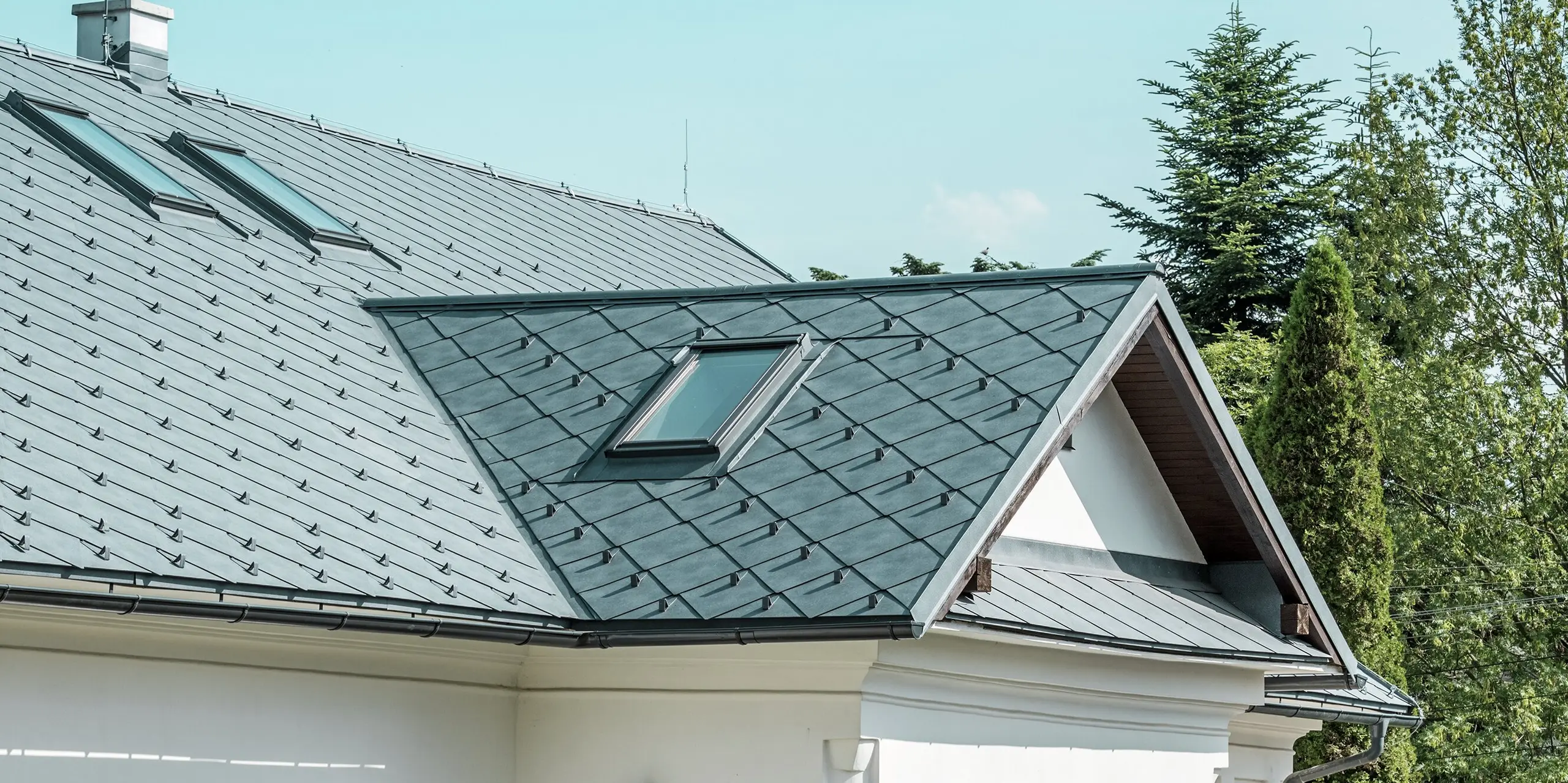 Das Bild zeigt einen detailreichen Ausschnitt des Handwerkerhauses in Klin, Slowakei. Die PREFA Dachraute 44 × 44 in P.10 Steingrau verleiht dem Dach ein modernes, ansprechendes Aussehen und fügt sich perfekt in die traditionelle Architektur des Gebäudes ein. Besonders auffällig sind die großzügigen Dachfenster, die Licht in den oberen Bereich des Hauses bringen und das Design zusätzlich auflockern. Das elegante Satteldach mit der Gaube über dem Eingangsbereich überzeugt mit Langlebigkeit, Leichtigkeit und Widerstandsfähigkeit, was es zur idealen Wahl für eine nachhaltige Sanierung macht.