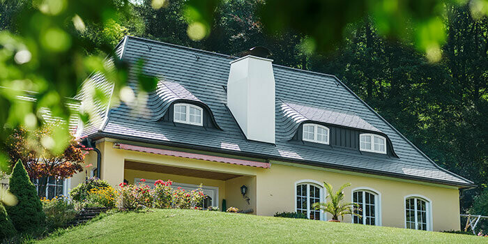 Maison individuelle avec toiture récemment rénovée à l’aide de bardeaux de toiture PREFA couleur anthracite, avec lucarnes arrondies (lucarne à joues galbées) et cheminée blanche.