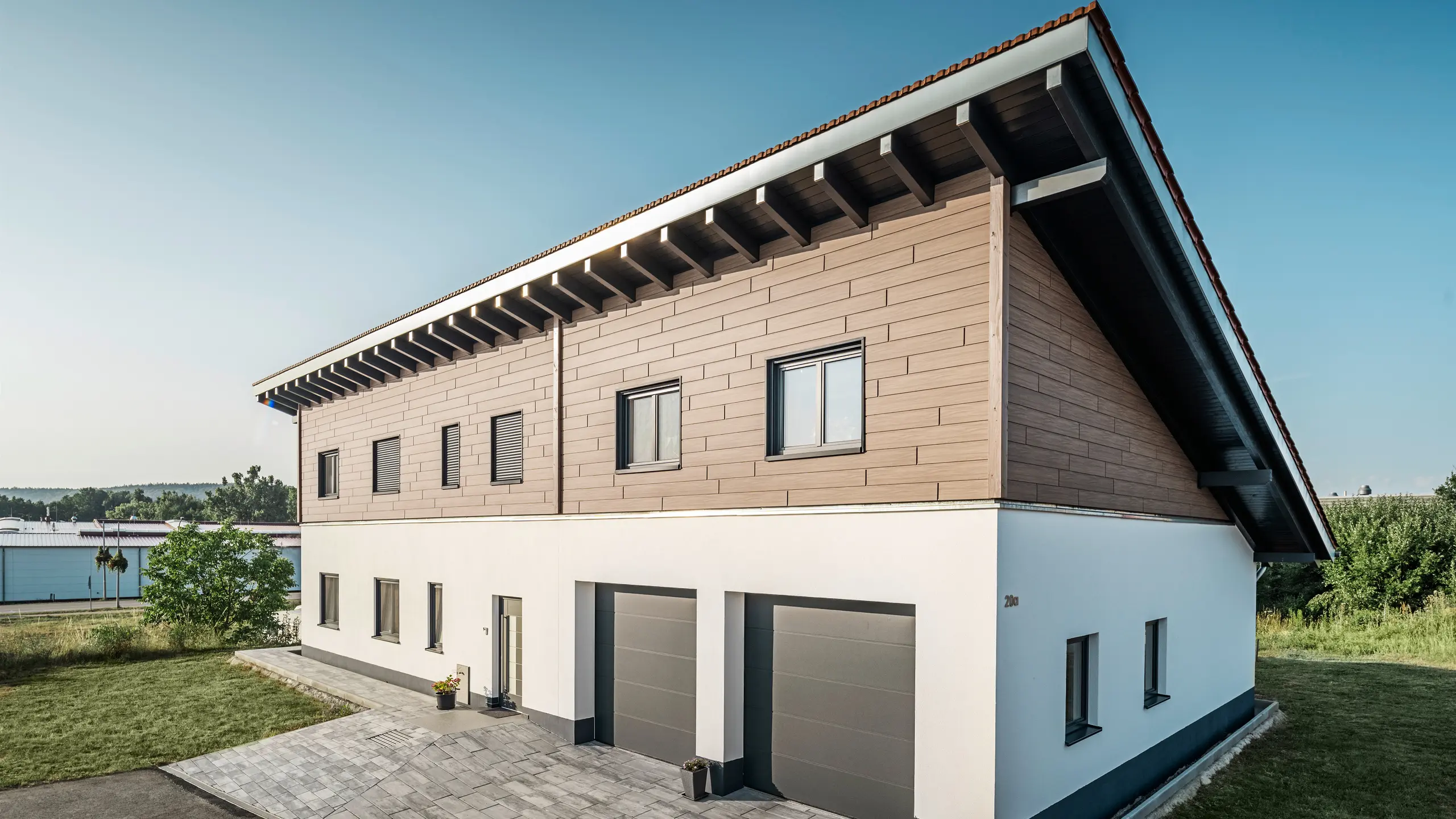 Blick von der Seite auf ein Einfamilienhaus mit Pultdach in Bruck. Das obere Stockwerk ist mit PREFA Sidings in Walnuss Braun ummantelt. Auf der rechten Seite des Bildes sind zwei Garagentore in Anthrazit zu sehen. In der Gebäudemitte befindet sich die Eingangstüre. Der Sockel ist in einem schlichten Weiß gehalten. 