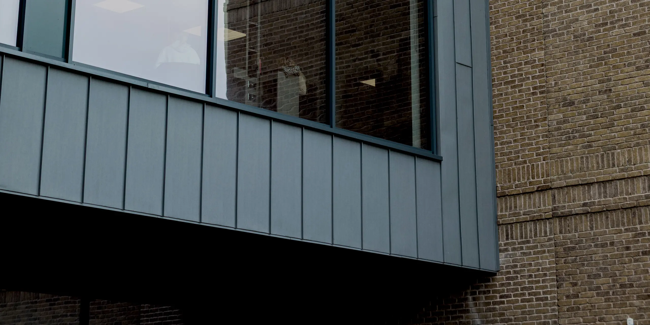 Une section remarquable de la façade du Northern Regional College de Ballymena présente une large façade vitrée, encadrée avec précision par des bacs en aluminium à joints debout PREFALZ gris quartz. La structure verticale de l'aluminium souligne la pureté des lignes et met élégamment en valeur le bâtiment en porte-à-faux. En arrière-plan, la maçonnerie de briques à la texture fine offre un contraste saisissant. La façade en aluminium PREFA, de haute qualité, impressionne par sa durabilité, sa résistance aux intempéries et son esthétique moderne et épurée, ce qui en fait un choix idéal pour l'architecture scolaire contemporaine.