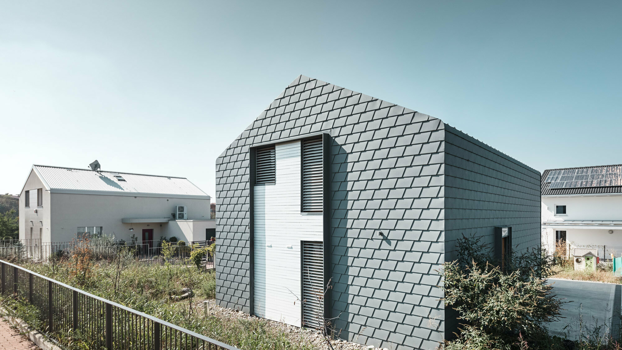 Vue de trois quarts de la maison et de son environnement, y compris deux autres maisons. Une rupture visuelle intervient sur la façade : il s’agit d’un lattage en bois blanc encadré de métal.