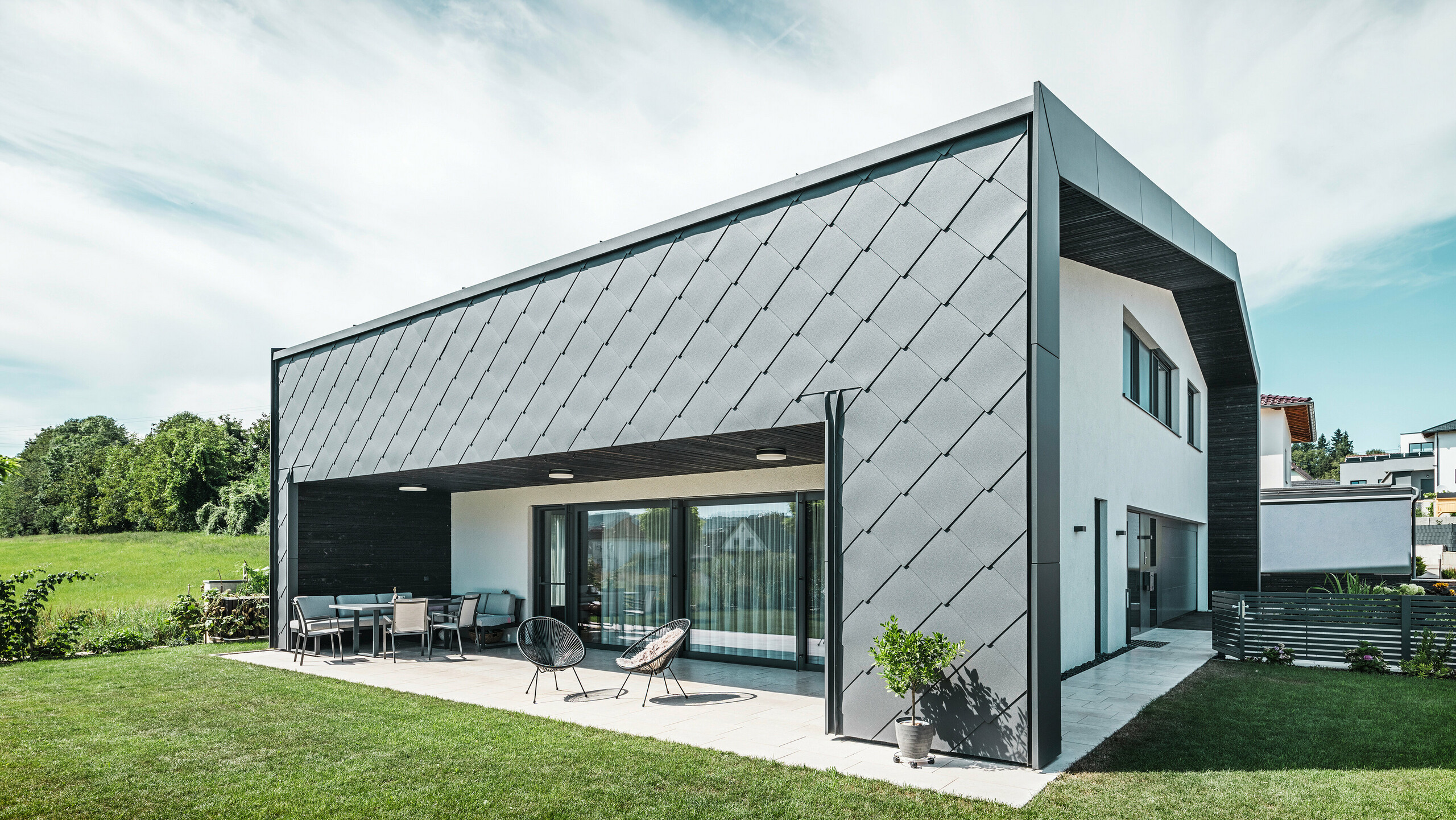 [Translate to Deutsch:] Gepflegter Gartenbereich eines Einfamilienhauses in Aschach. Das Gebäude ist stilvoll eingedeckt mit dunkelgrauen PREFA Aluminium-Wandrauten 44 × 44. Die überdachte Terrasse bietet einen gemütlichen Sitzplatz mit Blick auf die saftig grüne Rasenfläche. Die elegante Aluminiumverkleidung in Kombination mit weißen Putzflächen sorgt für eine moderne und witterungsbeständige Architektur. Große Fensterflächen lassen viel Tageslicht in den Innenraum und schaffen eine offene, lichtdurchflutete Atmosphäre.