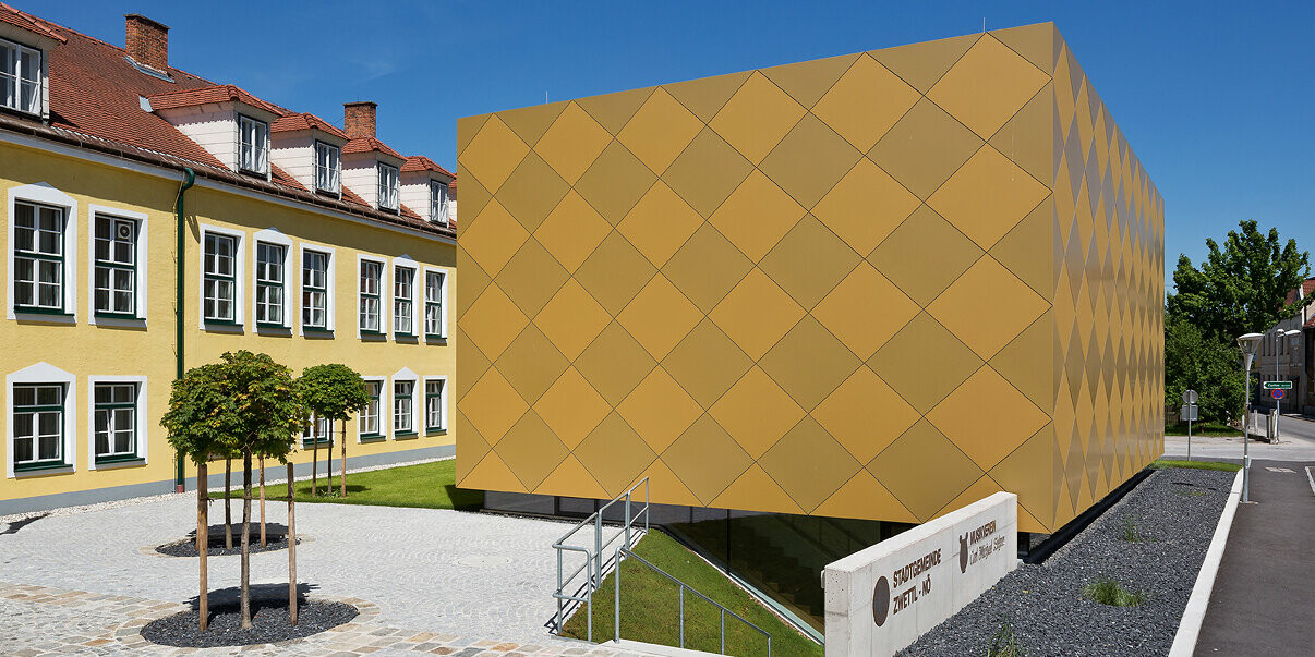 Blick aus der Distanz auf den Eingangsbereich des Musikvereins in Zwettl. Das quaderförmige Gebäude steht vor dem Stadtamt in Zwettl und sticht durch die markante Hülle aus PREFA Aluminium Verbundplatten hervor.