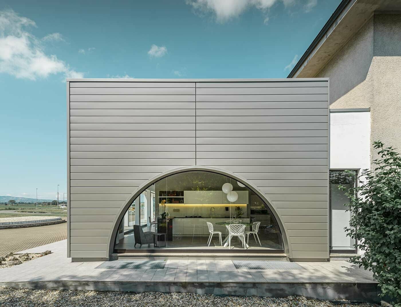 Extension avec toit plat et façade vitrée courbe, habillée de panneaux aluminium posés à l’horizontale : les Sidings PREFA couleur argent fumé.