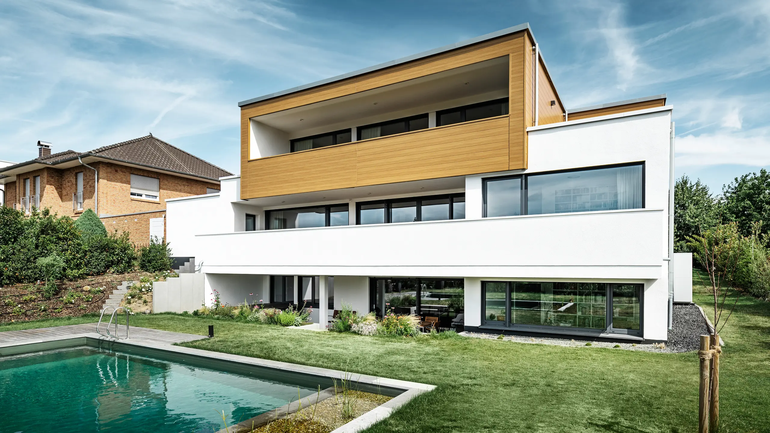 De achterkant van een modern vrijstaand huis in Usingen, Duitsland. Het groene gazon, een zwembad en een jonge tuinboom zijn onderaan de foto te zien. Het in het oog springende huis heeft een witte bouwbasis van een bepleisterde gevel, een aluminium gevel op de bovenverdieping van PREFA Sidings in eiken naturel en grote raamoppervlakken met een antracietkleurig kozijn. Van een afstand is het buurhuis met een bakstenen gevel aan de linkerkant van de foto te zien.  Vertaald met DeepL.com (gratis versie)