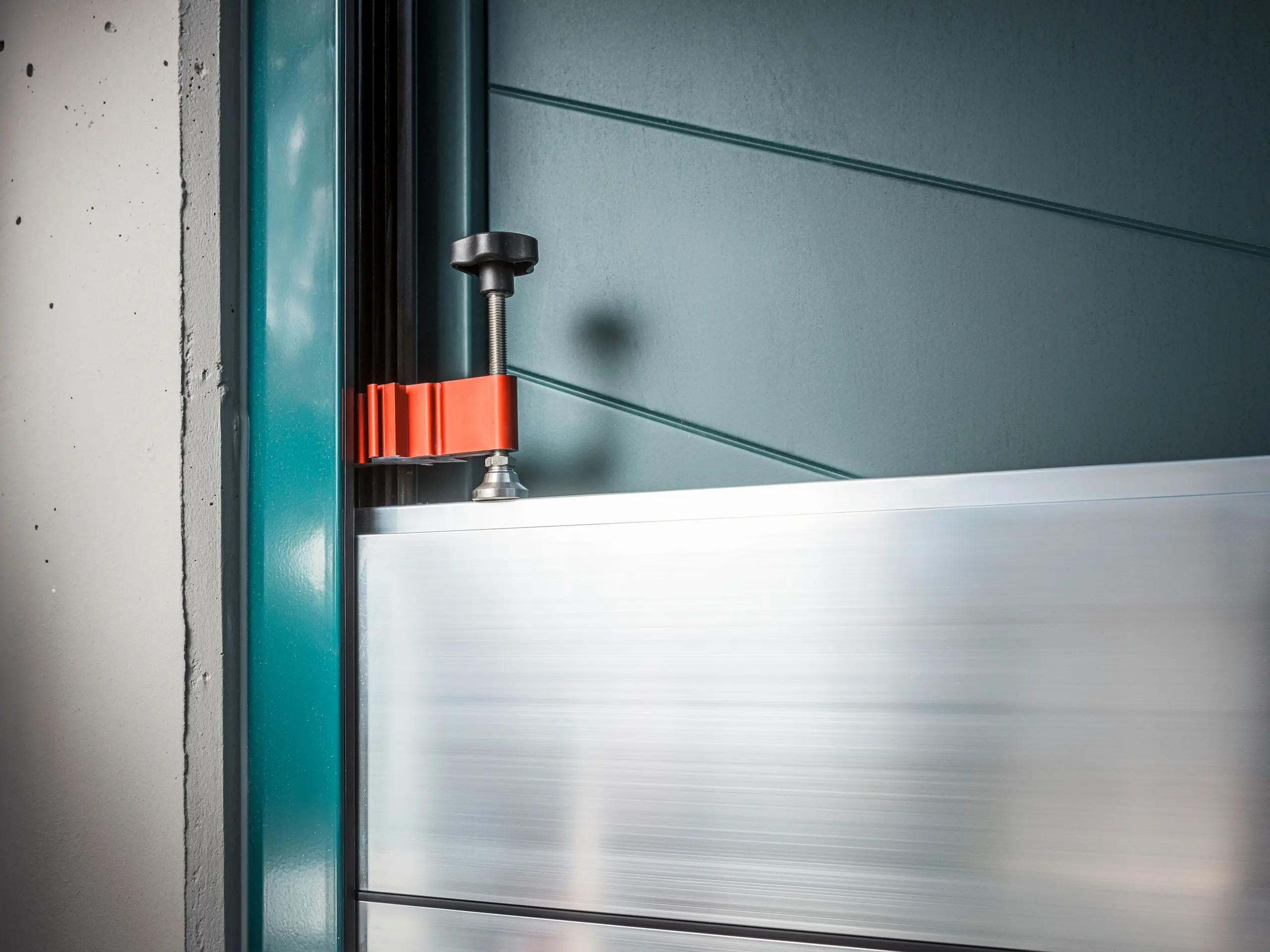 Close-up van het PREFA-overstromingsbeveiligingssysteem op een poortframe, bestaande uit aluminium dambalken bevestigd met een robuuste, rode klem (het klemstuk) voor een veilige bescherming tegen overstromingen.