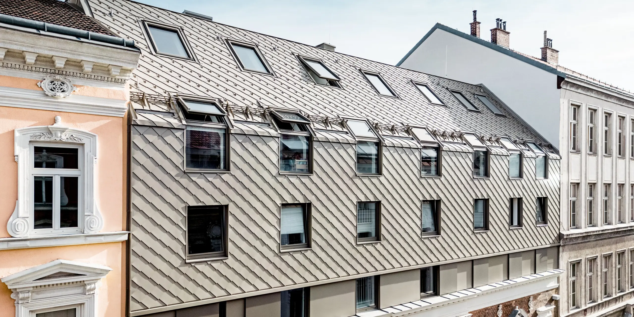 L'image révèle l'extension remarquable d'un immeuble d'habitation viennois, où des losanges 29x29 PREFA modernes dans la teinte bronze ornent l'étage supérieur. Ces losanges confèrent à l'architecture une touche élégante et contemporaine qui s'harmonise parfaitement avec la façade historique. Les lucarnes, particulièrement remarquables, animent la toiture et créent une structure intéressante. Le contraste entre le mur de briques classique et les losanges en aluminium modernes souligne la fusion réussie de tradition et d'innovation dans l'architecture viennoise.