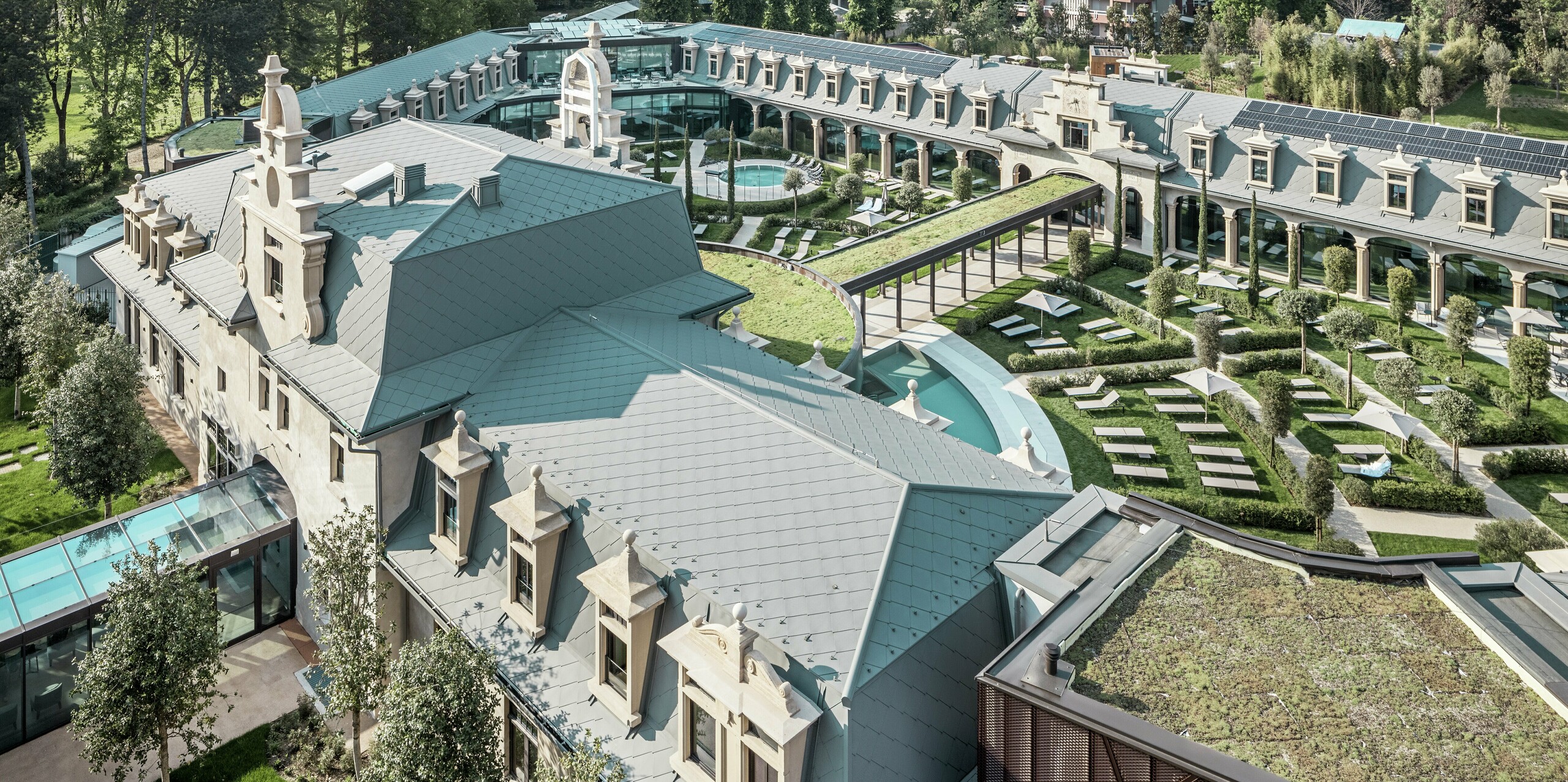Vue panoramique du complexe thermal « De Montel – Terme Milano », entièrement rénové, avec ses vastes jardins, ses allées panoramiques et ses espaces détente autour de ses nombreuses piscines thermales. Les toitures sont entièrement recouvertes de losanges PREFA 44 × 44 P.10 gris souris, complétées par des modules photovoltaïques parfaitement intégrés et des espaces paysagers. Architecture historique et matériaux durables se conjuguent pour créer un espace de loisirs urbain innovant.