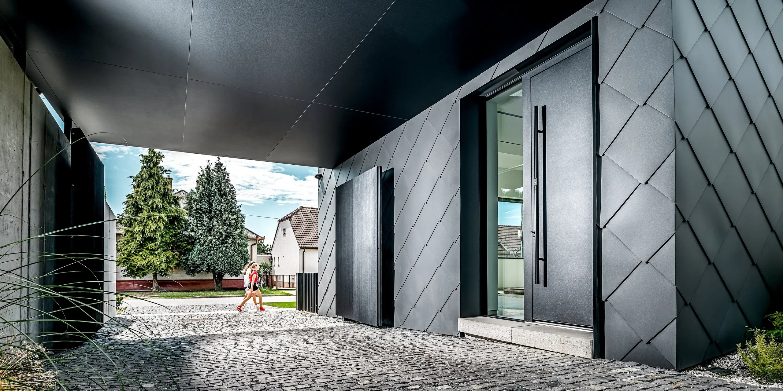 Cette image présente l'entrée et l'allée d'une maison individuelle moderne à Trnava-Modranka, dont la façade remarquable est entièrement revêtue de losanges de façade 44x44 PREFA, P.10 gris sombre. Le motif précis des losanges confère au bâtiment une allure élégante et futuriste, tout en soulignant la pureté des lignes architecturales. Ces produits en aluminium résistant offrent une protection durable contre les intempéries et une apparence haut de gamme et pérenne. À l'arrière-plan, la végétation luxuriante crée un contraste naturel avec l'enveloppe sombre du bâtiment.