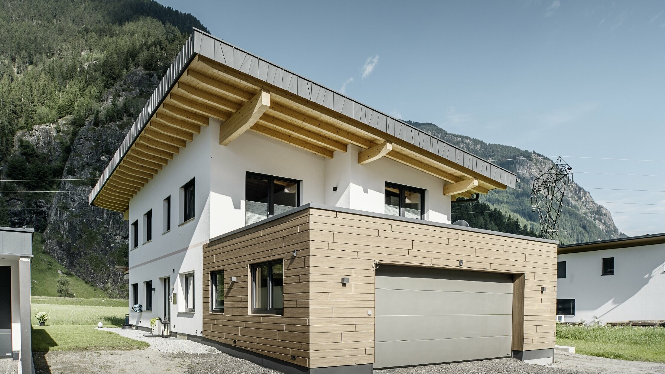 Moderne eengezinswoning met gevelrenovatie: vernieuwde buitengevel met horizontaal geplaatste gevelbekleding in houtlook, gecombineerd met wit gepleisterde vlakken, geïntegreerde garagebekleding en eigentijdse architectuur in een alpiene omgeving.