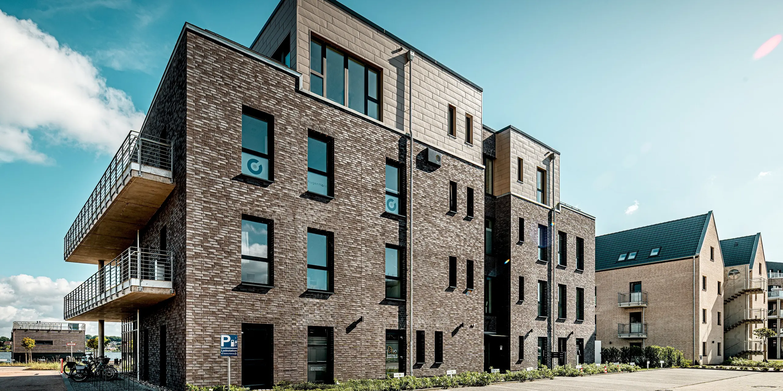 L'image présente un bâtiment du complexe résidentiel « Hafen zur Freiheit » (Port vers la liberté), dont l'allure résolument moderne est due à l'alliance d'une façade en briques sombres et des panneaux Siding.X PREFA couleur bronze du dernier étage. Les surfaces en aluminium à la structure apparente créent un contraste élégant avec l'aspect traditionnel de la brique et confèrent au bâtiment une allure dynamique et haut de gamme. De larges fenêtres et des balcons latéraux soulignent l'architecture ouverte. Grâce à la robustesse et à la résistance aux intempéries des éléments PREFA, la façade bénéficie d'une protection permanente et d'une stabilité visuelle optimale, même en milieu maritime.