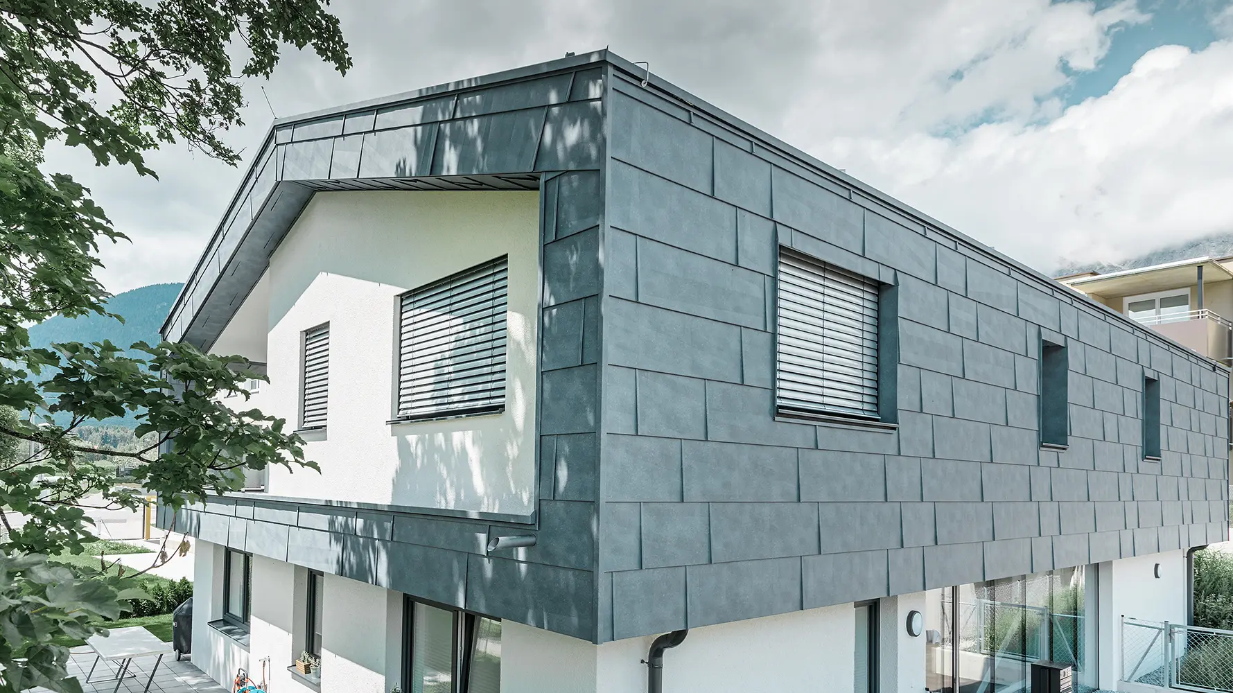 Maison individuelle moderne sans avancée de toit. L’étage supérieur a été recouvert de panneaux de façade FX.12 PREFA couleur gris pierre et le reste de la façade a été crépi en blanc.