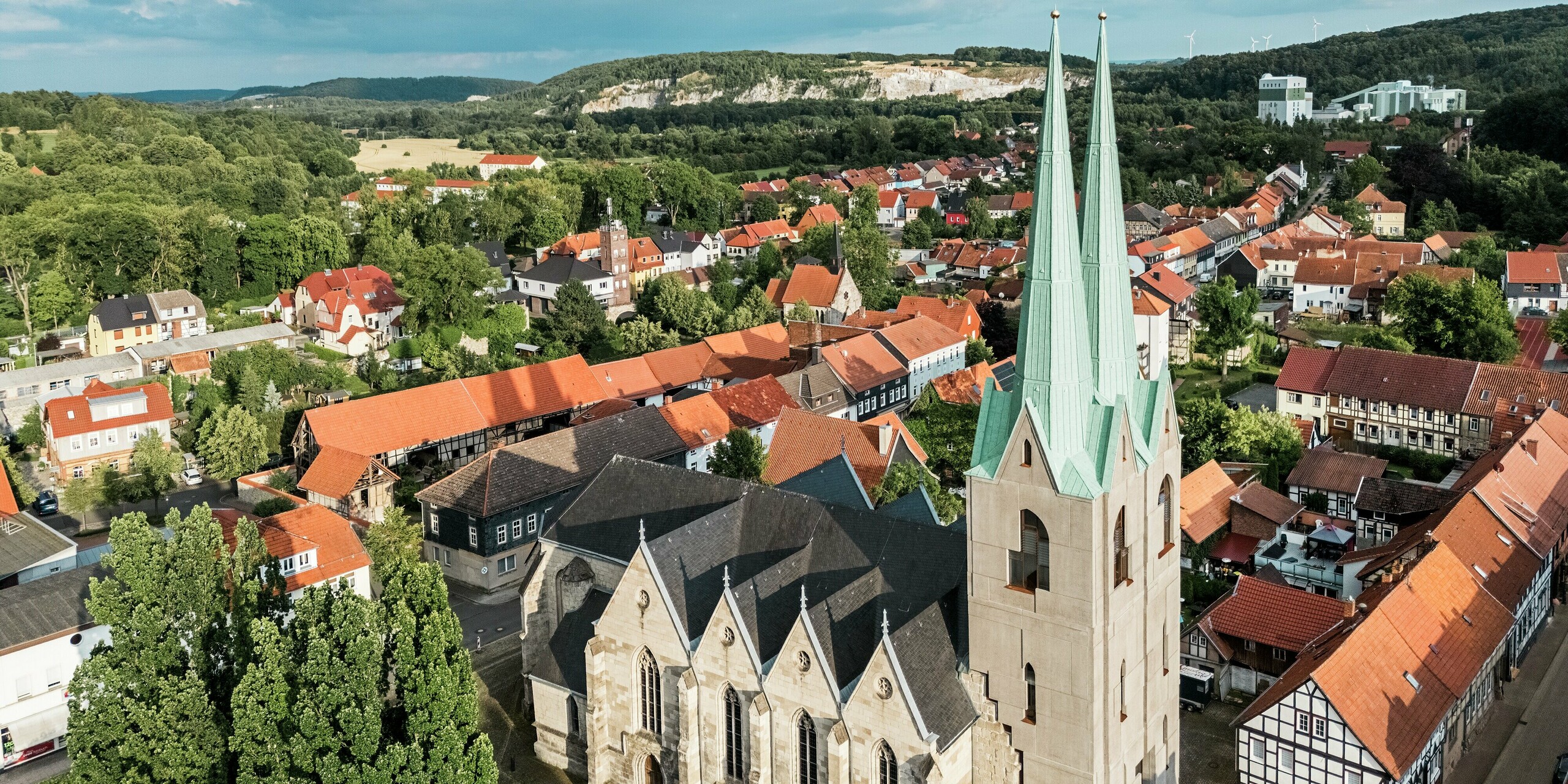Drohnenaufnahme der Stadtkirche St. Johannis in Ellrich, Thüringen, mit markanten Turmspitzen aus PREFALZ in P.10 Patinagrün. Das moderne Aluminiumdach setzt einen klaren architektonischen Akzent im historischen Ortskern. Umgeben von roten Ziegeldächern und Fachwerkhäusern, fügt sich das langlebige, wartungsfreie Dachmaterial harmonisch und gleichzeitig selbstbewusst ins Stadtbild ein.