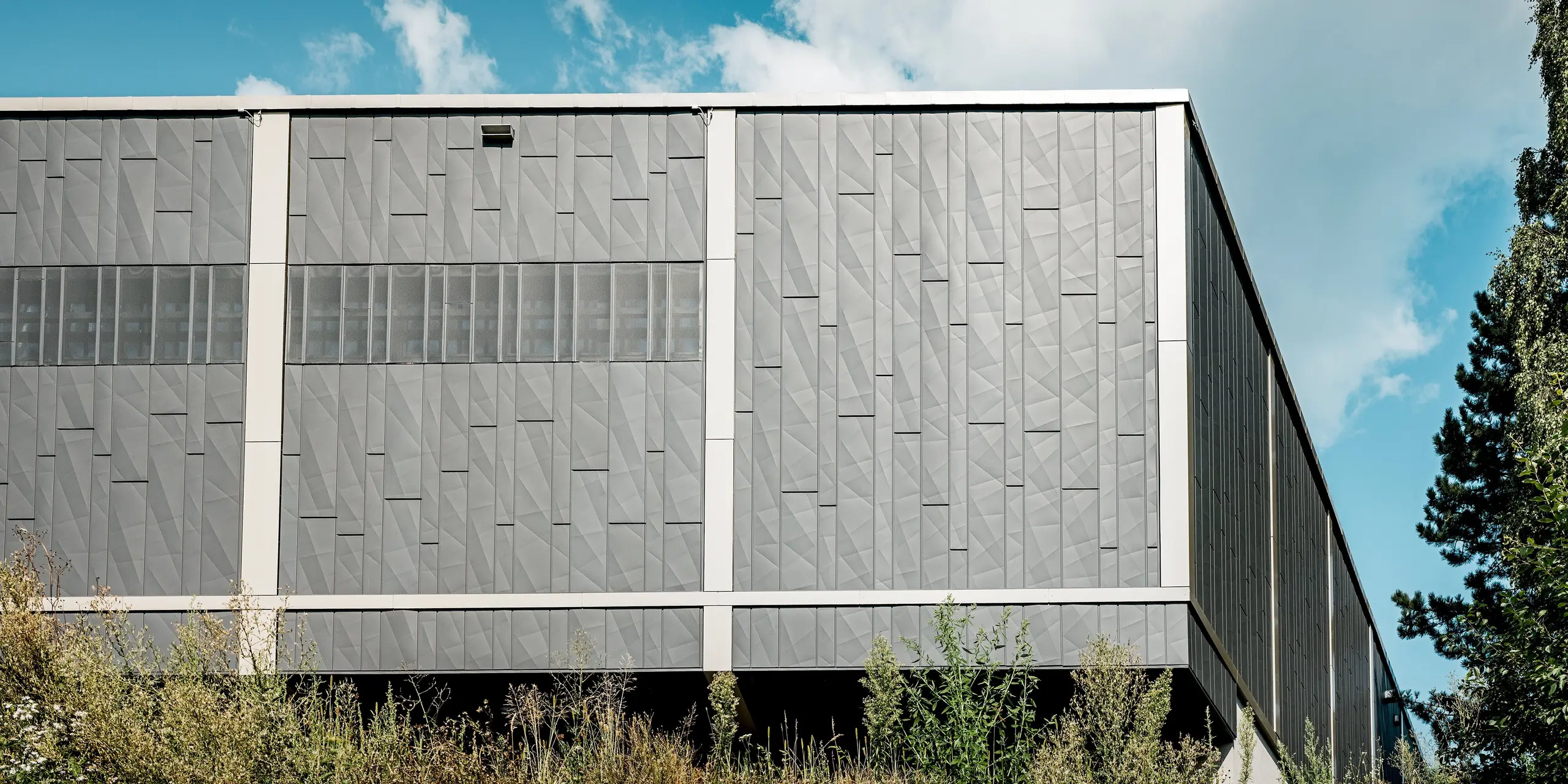 L'image présente la façade arrière du centre commercial de Rohrbach, dotée d'une façade métallique moderne composée de panneaux Siding.X PREFA anthracite. La structure géométrique des panneaux crée une texture dynamique, conférant au bâtiment une esthétique contemporaine et saisissante. Les bandes horizontales perforées attirent particulièrement le regard, offrant un intérêt visuel tout en assurant une ventilation ciblée et une transmission optimale de la lumière. La façade s'intègre harmonieusement à son environnement naturel, le jeu entre les riches nuances de gris et la végétation luxuriante créant un lien captivant entre architecture et paysage.