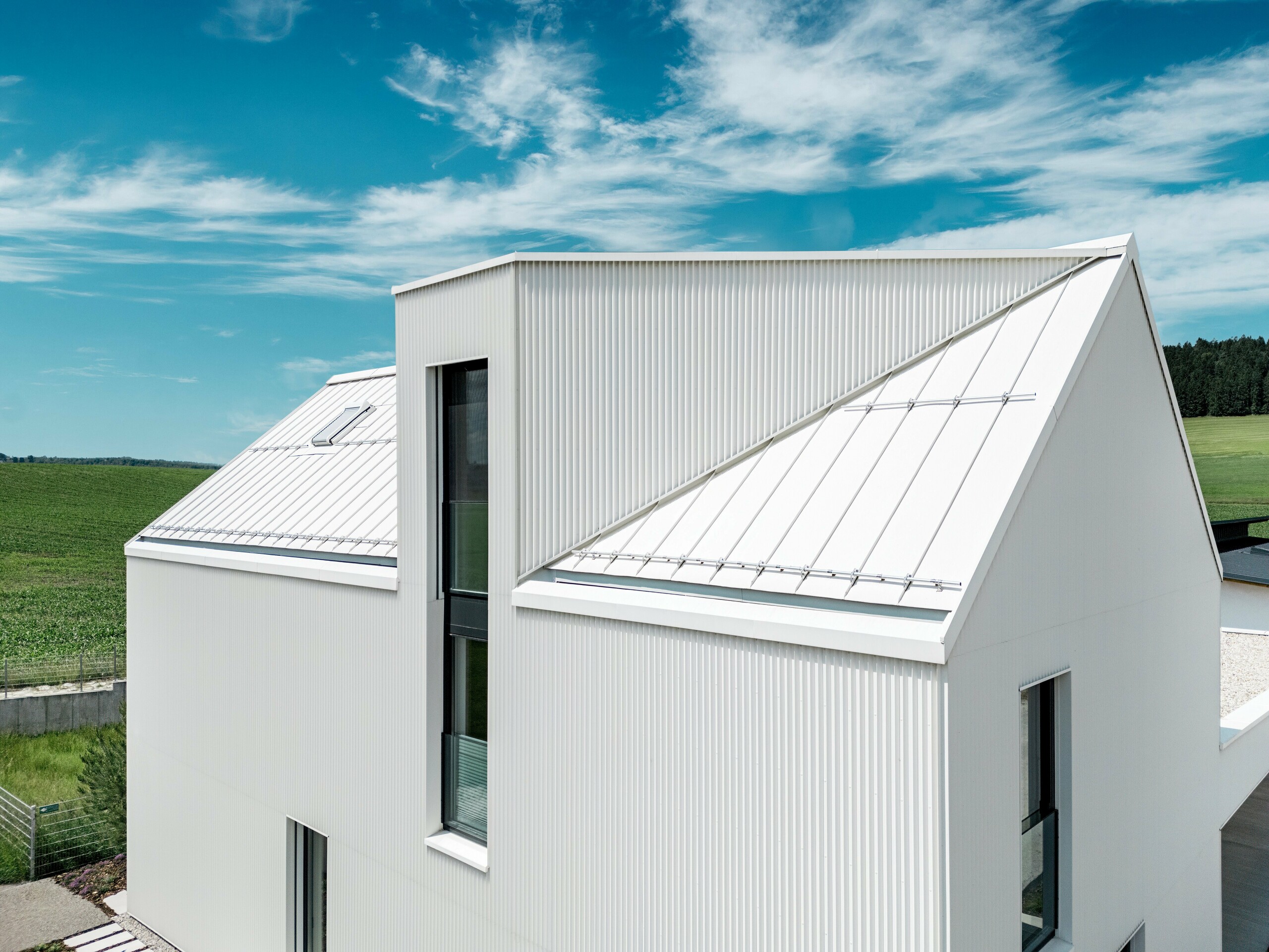 Das Bild zeigt das steile, markante Satteldach dieses Einfamilienhauses in Freistadt, das mit PREFALZ in der Farbe Reinweiß verkleidet ist. Die präzise gefaltete Fassade und das Dach sind nahtlos miteinander verbunden, was eine elegante, klare Linienführung erzeugt. Das verwendete PREFALZ für Dach und Fassade gewährleistet nicht nur eine ästhetische Beständigkeit, sondern auch eine hohe Witterungsbeständigkeit. Dadurch wird dem Gebäude langfristige Langlebigkeit und Pflegeleichtigkeit verliehen. Das Zusammenspiel von modernem Design und funktionalen Materialien schafft eine harmonische, zeitgemäße Architektur, die sowohl praktisch als auch optisch überzeugt. Besonders ins Auge fällt die Gaube mit dem schrägen Verlauf und dem über zwei Etagen ragenden Fenster.