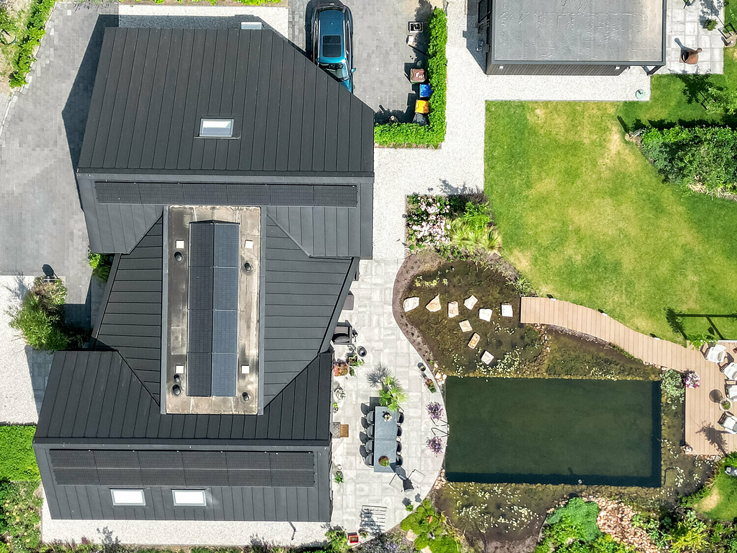 Luftaufnahme eines modernen Einfamilienhauses mit einem komplexen Dachsystem aus schwarzem Prefalz, Solarmodulen und mehreren Gauben. Das Anwesen verfügt über einen gepflegten Garten mit Rasenflächen, einem großen Teich, einem Holzsteg und einer Sitzgruppe. Gepflasterte Wege und eine Einfahrt vervollständigen das Bild der ordentlich angelegten Außenanlage, die das Haus umgibt.
