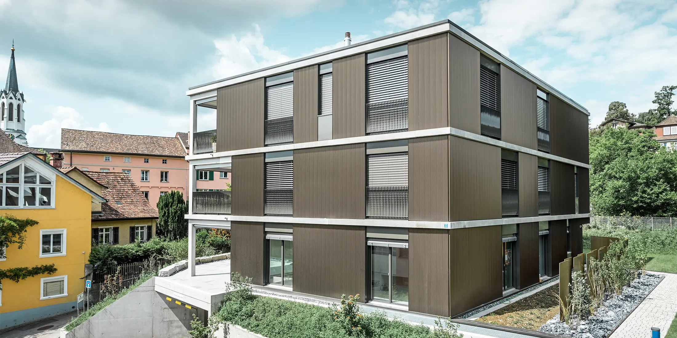 Die markante Aluminiumfassade dieses Mehrfamilienhauses in Schaffhausen ist mit dem eleganten PREFA Zackenprofil in der Farbe Colinal ausgestattet. Durch die vertikale Anordnung der stranggepressten Aluminiumprofile wirkt das Gebäude optisch kompakter und die Fassadenstruktur erhält eine harmonische Balance. Das edle Design fügt sich geschmackvoll in die Umgebung ein und verleiht dem Gebäude eine moderne Eleganz, die sowohl optisch ansprechend als auch funktional ist. Die Perspektive zeigt das Mehrfamilienhaus von der Vorderseite mit der Garageneinfahrt. Im Hintergrund sind Bäume, weitere Gebäude und ein Kirchturm zu erkennen.