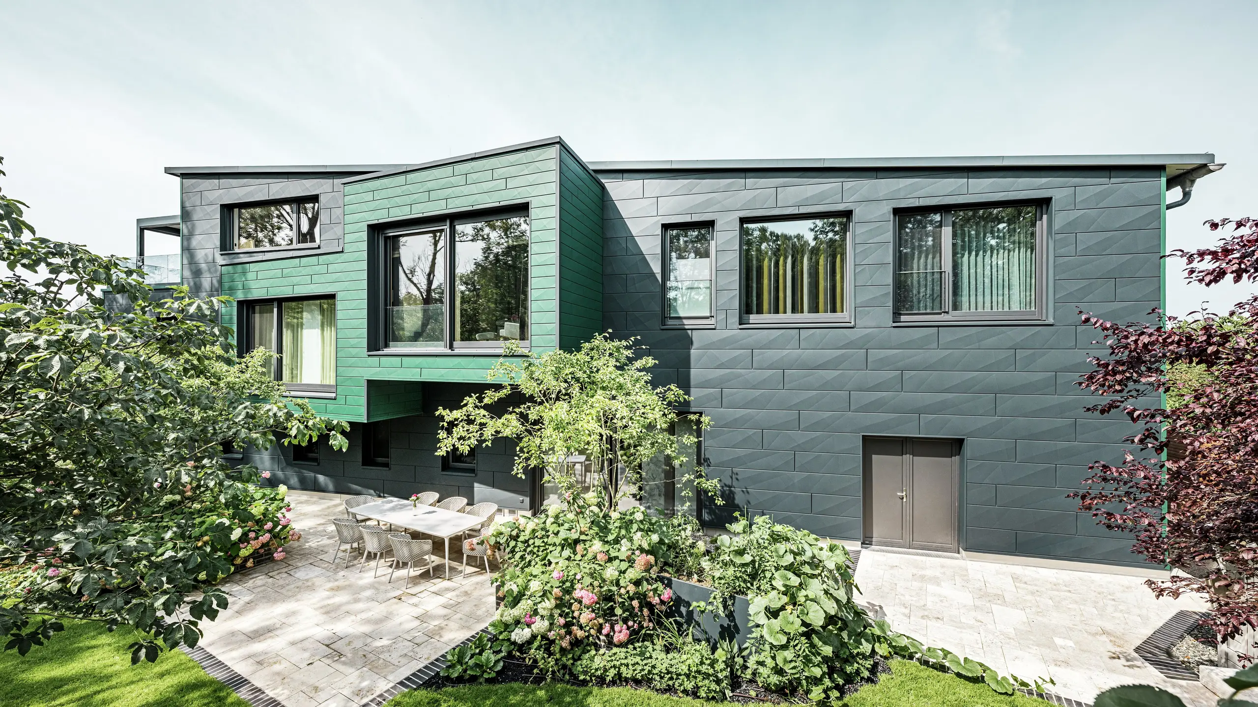 Das Einfamilienhaus in Türkheim beeindruckt durch seine außergewöhnliche Fassade, die mit PREFA Siding X-Paneelen in den Farben Anthrazit und Moosgrün verkleidet ist. Die klaren Linien und die markante Farbwahl der Aluminiumpaneele verleihen dem Gebäude eine moderne und elegante Ausstrahlung. Im Vordergrund ist der großzügige Gartenbereich mit einer stilvollen Terrasse und einem Esstisch mit modernen Stühlen zu sehen. Bunte Blumenbeete und gesunde Pflanzen sorgen für einen lebendigen, grünen Kontrast zur robusten Fassade. Die langlebige und pflegeleichte Aluminiumfassade schützt das Gebäude und sorgt für zeitlose Schönheit.