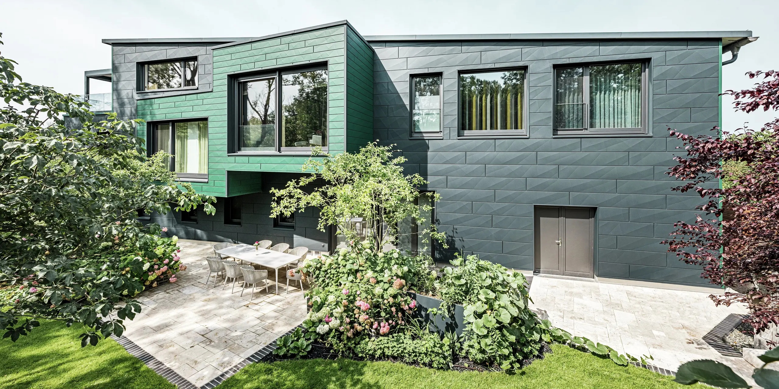 De eengezinswoning in Türkheim maakt indruk met zijn bijzondere gevel, die is bekleed met PREFA Siding X-panelen in de kleuren antraciet en mosgroen. De strakke lijnen en de opvallende kleurkeuze van de aluminium panelen geven het gebouw een moderne en elegante uitstraling. Op de voorgrond is de royale tuin te zien met een stijlvol terras en een eettafel met moderne stoelen. Kleurrijke bloembedden en gezonde planten zorgen voor een levendig, groen contrast met de robuuste gevel. De duurzame en onderhoudsvriendelijke aluminium gevel beschermt het gebouw en zorgt voor tijdloze schoonheid.