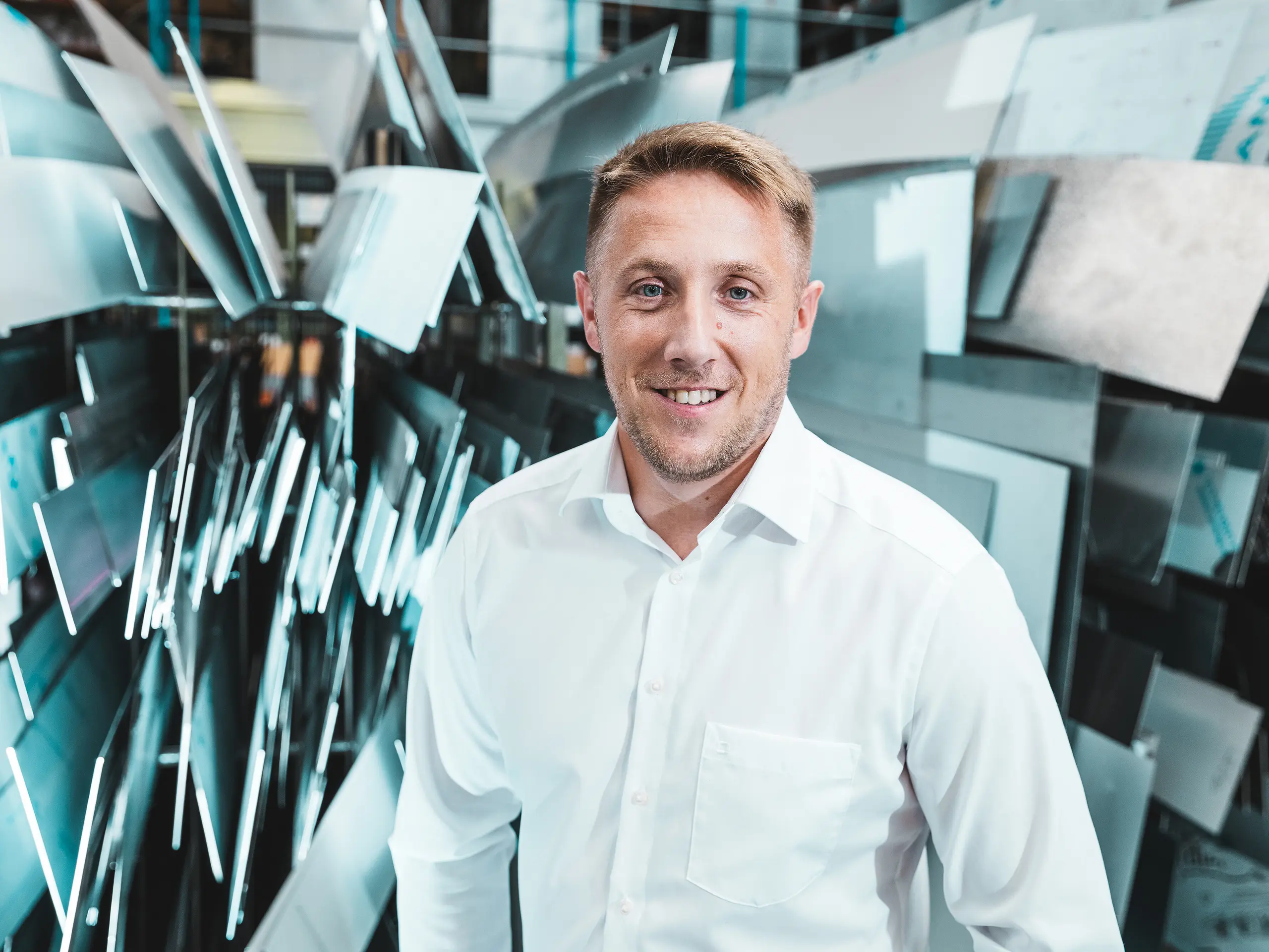Confiant et affable, Marcel Schmidt, chef de projet chez ASA Schüßler, entreprise spécialisée dans les toitures métalliques, se tient devant un impressionnant mur composé d'éléments en aluminium de formes variées. Les panneaux métalliques, agencés avec dynamisme, reflètent efficacement la lumière et symbolisent l'innovation, la précision et la qualité du travail des métaux. Cet homme en chemise blanche incarne la compétence et l'excellence de la production moderne. À l'arrière-plan, on découvre la diversité des possibilités de transformation de l'aluminium : des systèmes de toiture et de façade aux solutions spéciales sur mesure.