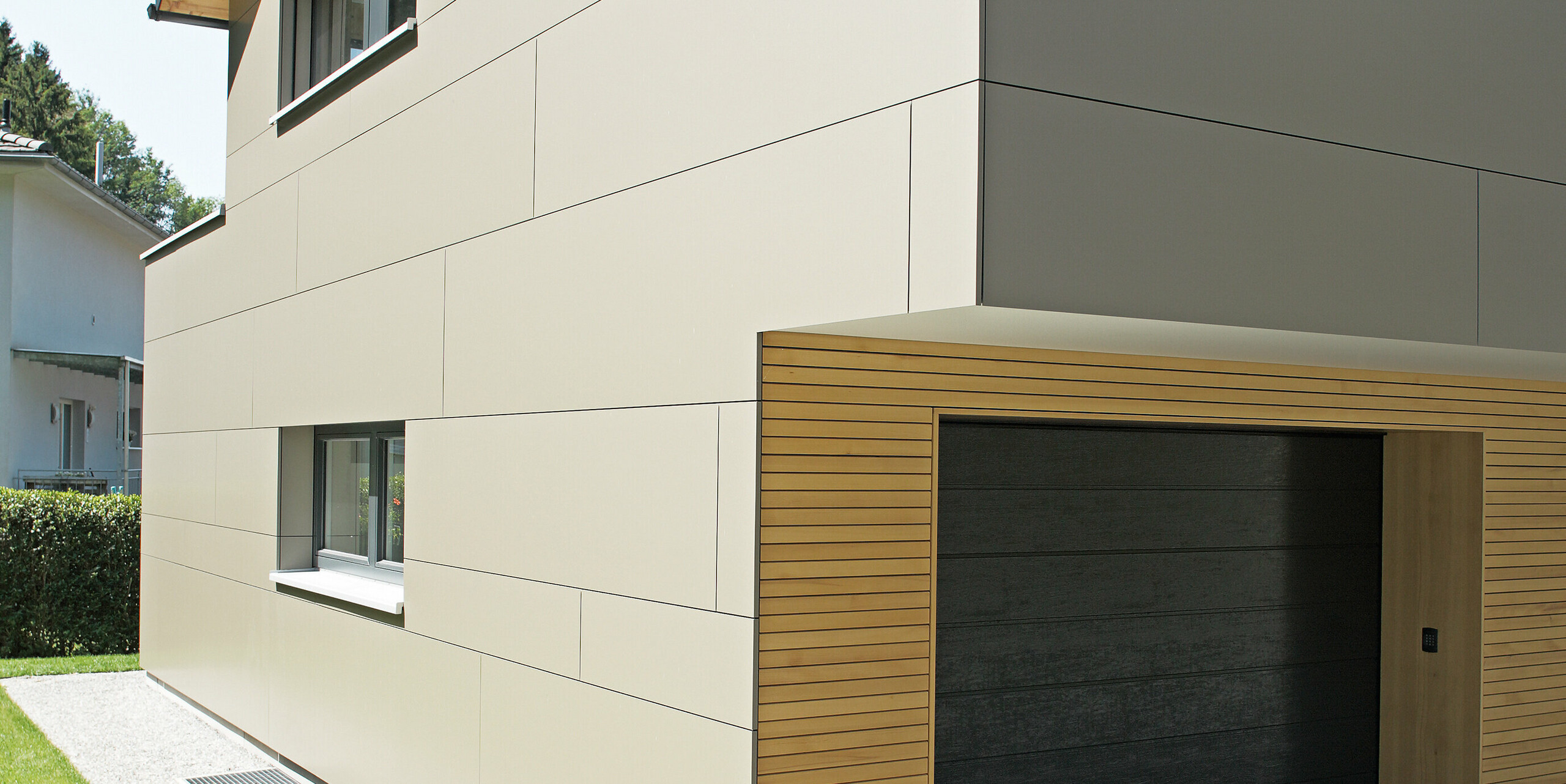 Moderne Architektur eines Einfamilienhauses in Hohenems, gezeigt an der Ecke mit Garage. Die Fassade besteht aus PREFA Aluminium Verbundplatten in der Farbe Bronzemetallic, die eine klare, minimalistische Ästhetik bieten. Die warmen Holzakzente um die Garage setzen einen natürlichen Kontrast zur metallischen Fassade, während das schräge Dach mit den dunklen PREFA Dachplatten in P.10 Anthrazit den konstruktiven Schutz und das durchdachte Design hervorhebt.