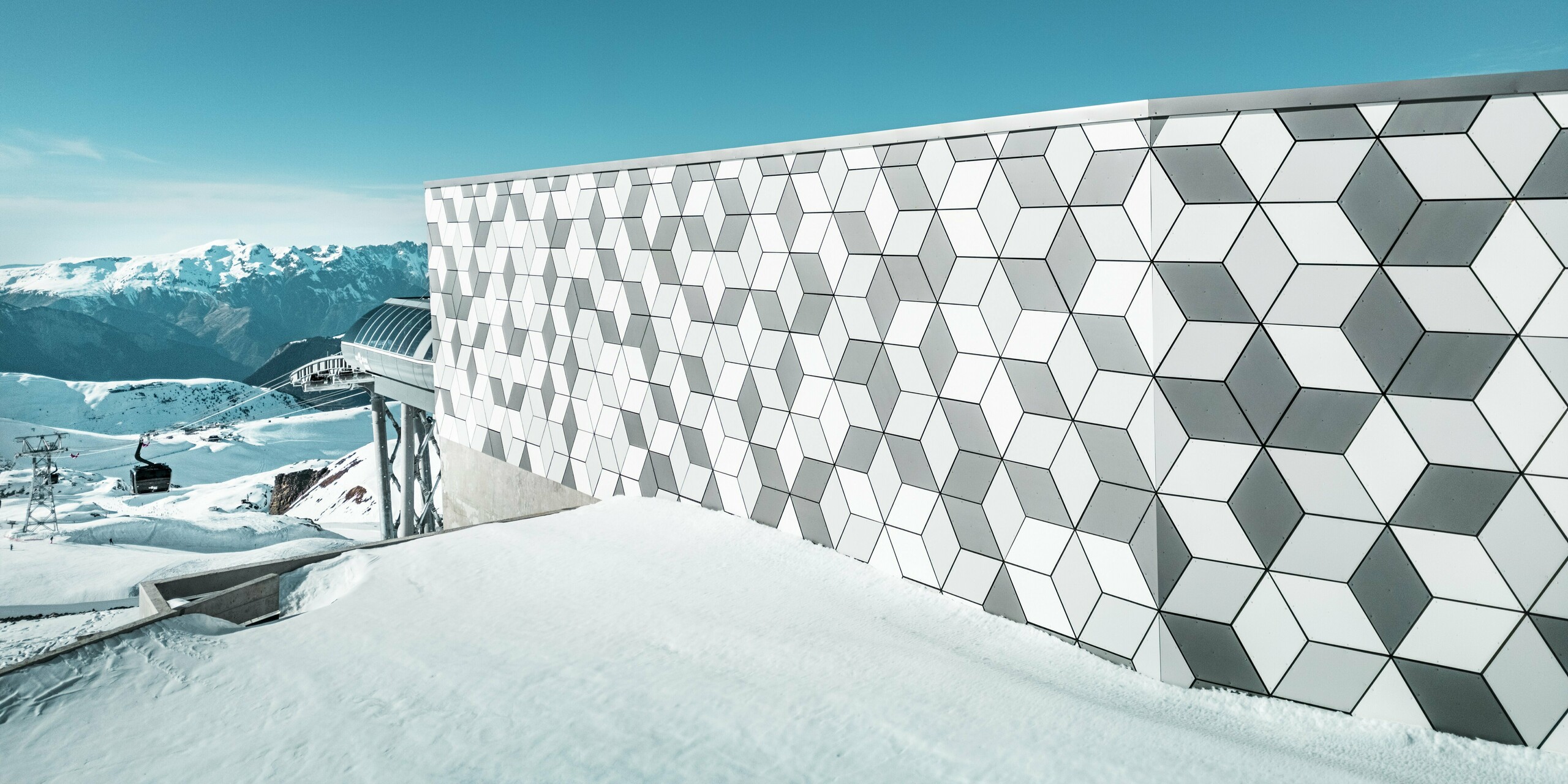 Photo de la façade en aluminium de la station de téléphérique des Deux Alpes : la façade imposante en panneaux composites en aluminium PREFABOND impressionne par son motif géométrique en losanges. L'harmonieuse combinaison de blanc et de gris crée un look moderne qui s'intègre harmonieusement dans le majestueux paysage montagneux. Derrière la façade, on aperçoit le ciel bleu éclatant et le majestueux panorama montagneux.