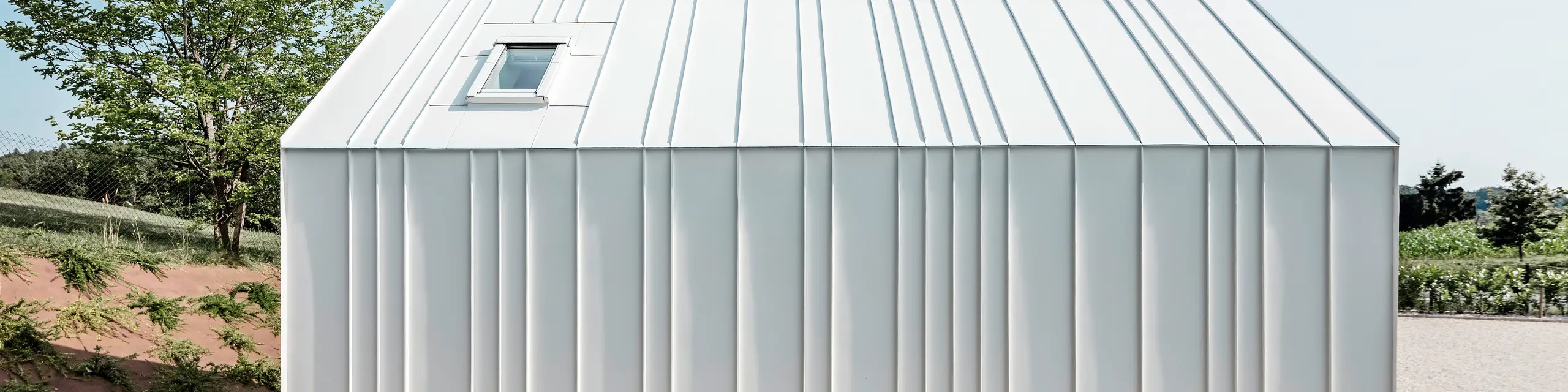 Vue latérale de l'une des petites maisons « Pri Momi » à Kančevci, en Slovénie. La micro-maison est recouverte d'un élégant toit à joints debout fabriqué à partir du système de toiture et de façade durable PREFALZ. Le revêtement en aluminium de haute qualité est de couleur blanc pur P.10. L'image montre les plis exécutés avec précision et les lignes élégantes de la coque en aluminium blanc, qui confèrent au bâtiment un aspect moderne et minimaliste. L'environnement naturel et le design clair et simple s'harmonisent parfaitement.